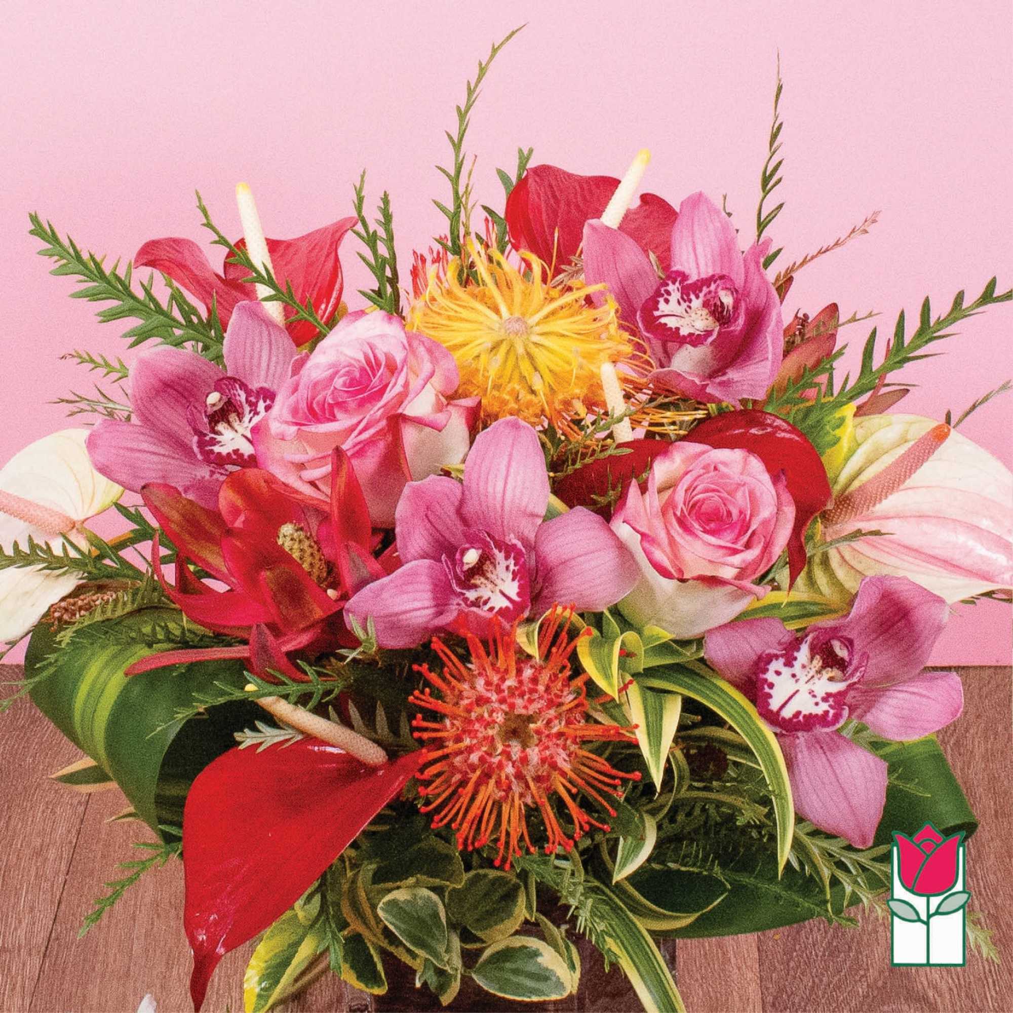 Low tropical arrangement with pink roses, orchids, red anthurium, and yellow protea in a glass vase.