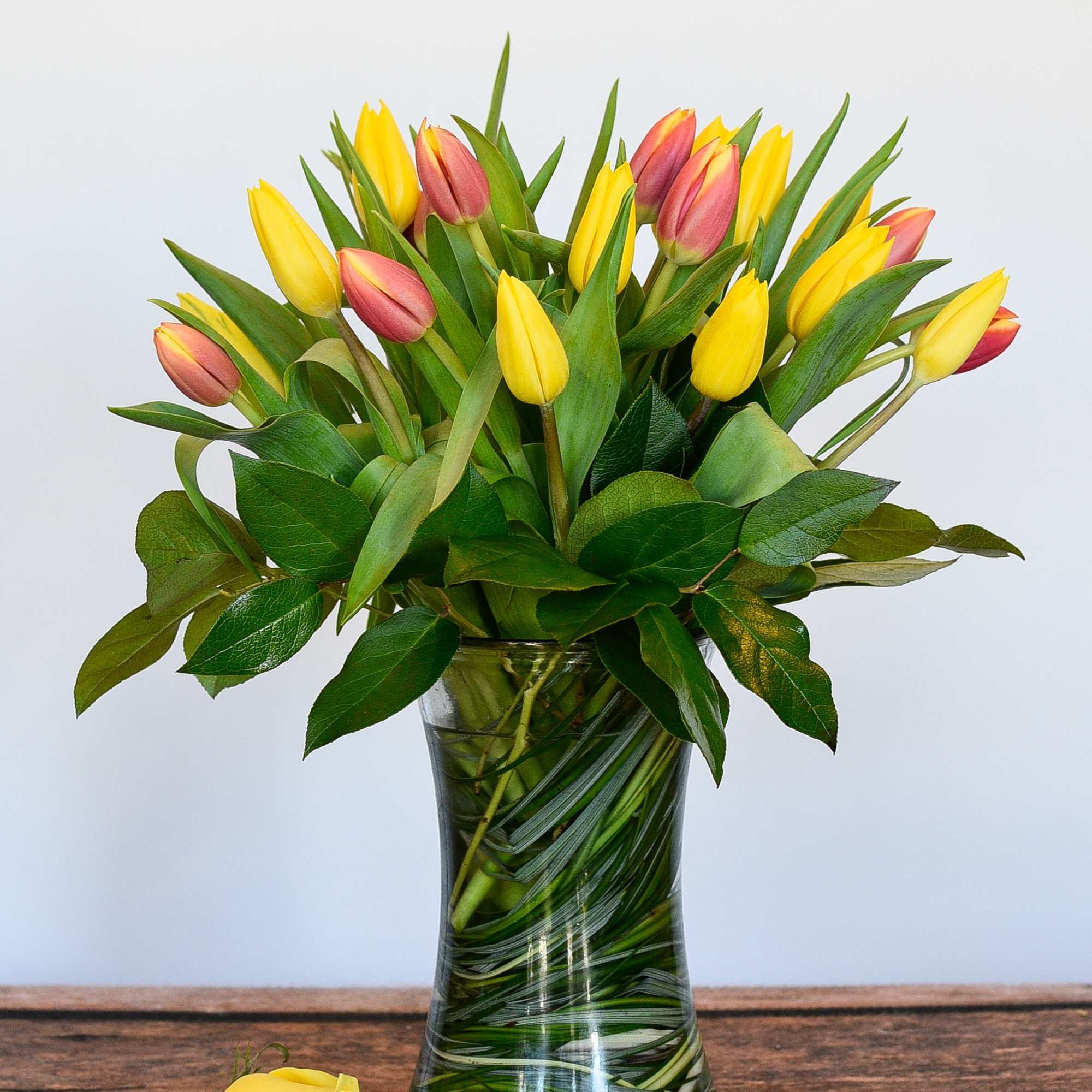 Yellow and pink tulips in a clear glass vase with a single yellow rose lying beside it