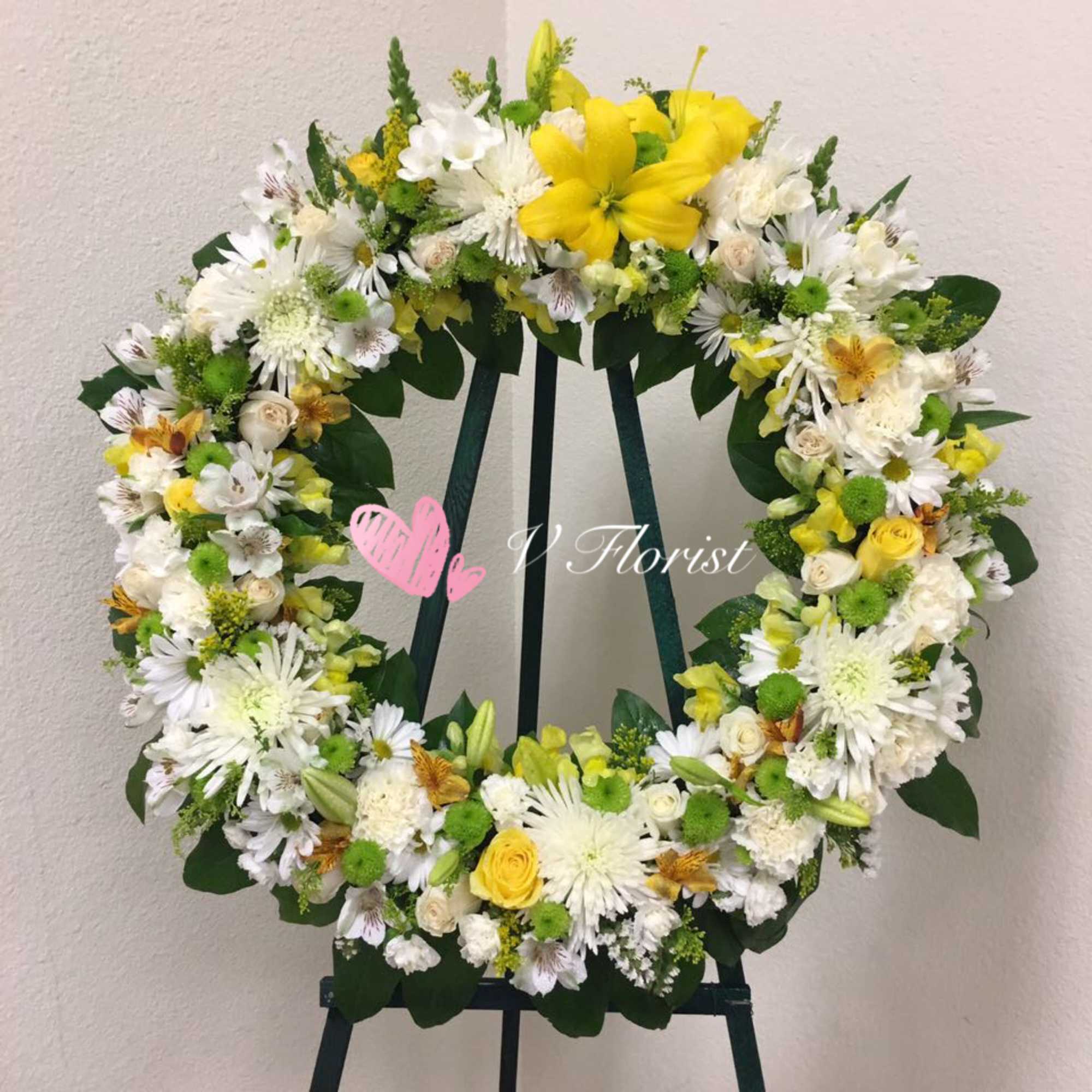 Standing wreath of yellow lilies, roses, and white flowers on a green easel.