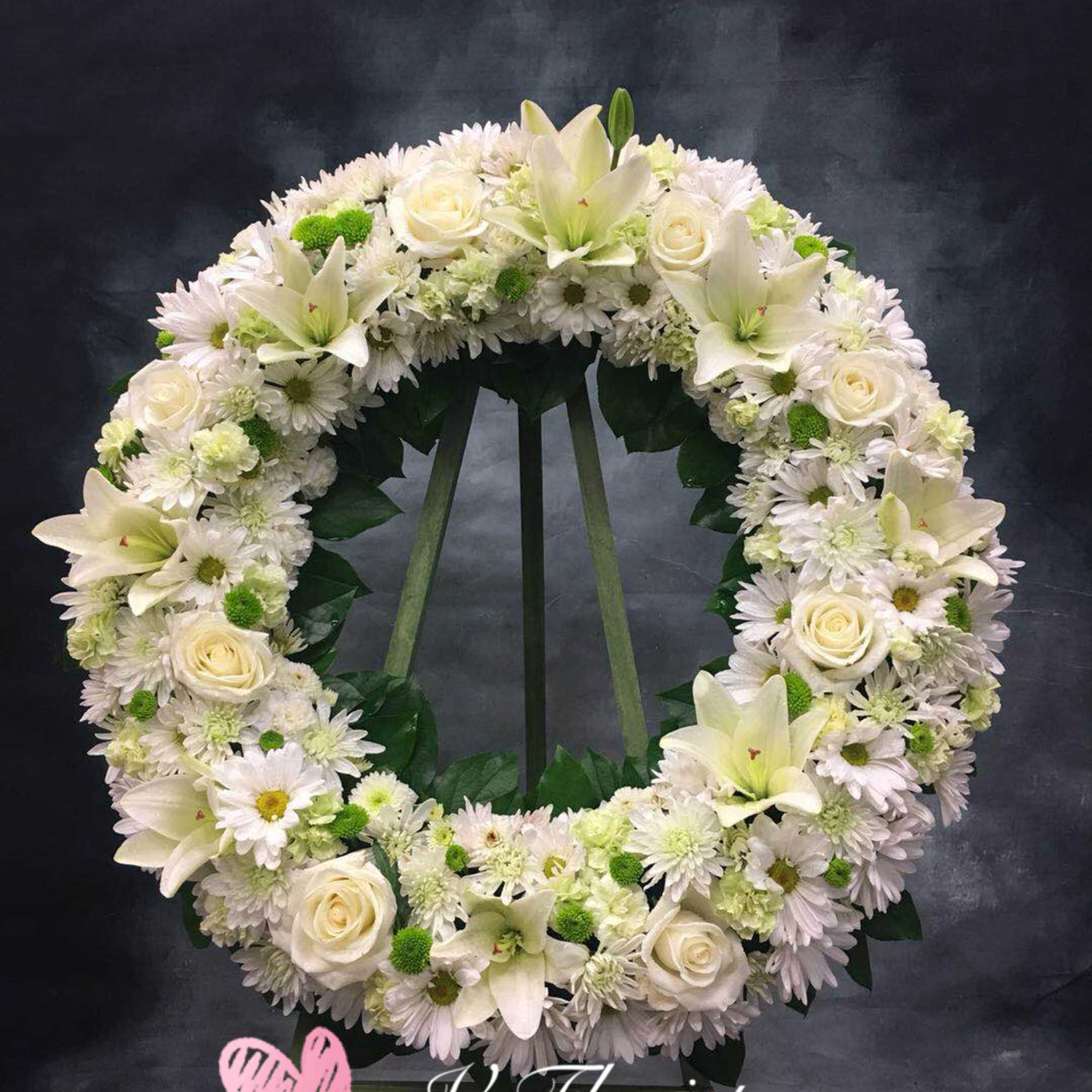 Large white and green standing wreath with roses, lilies, and daisies on a tripod stand.