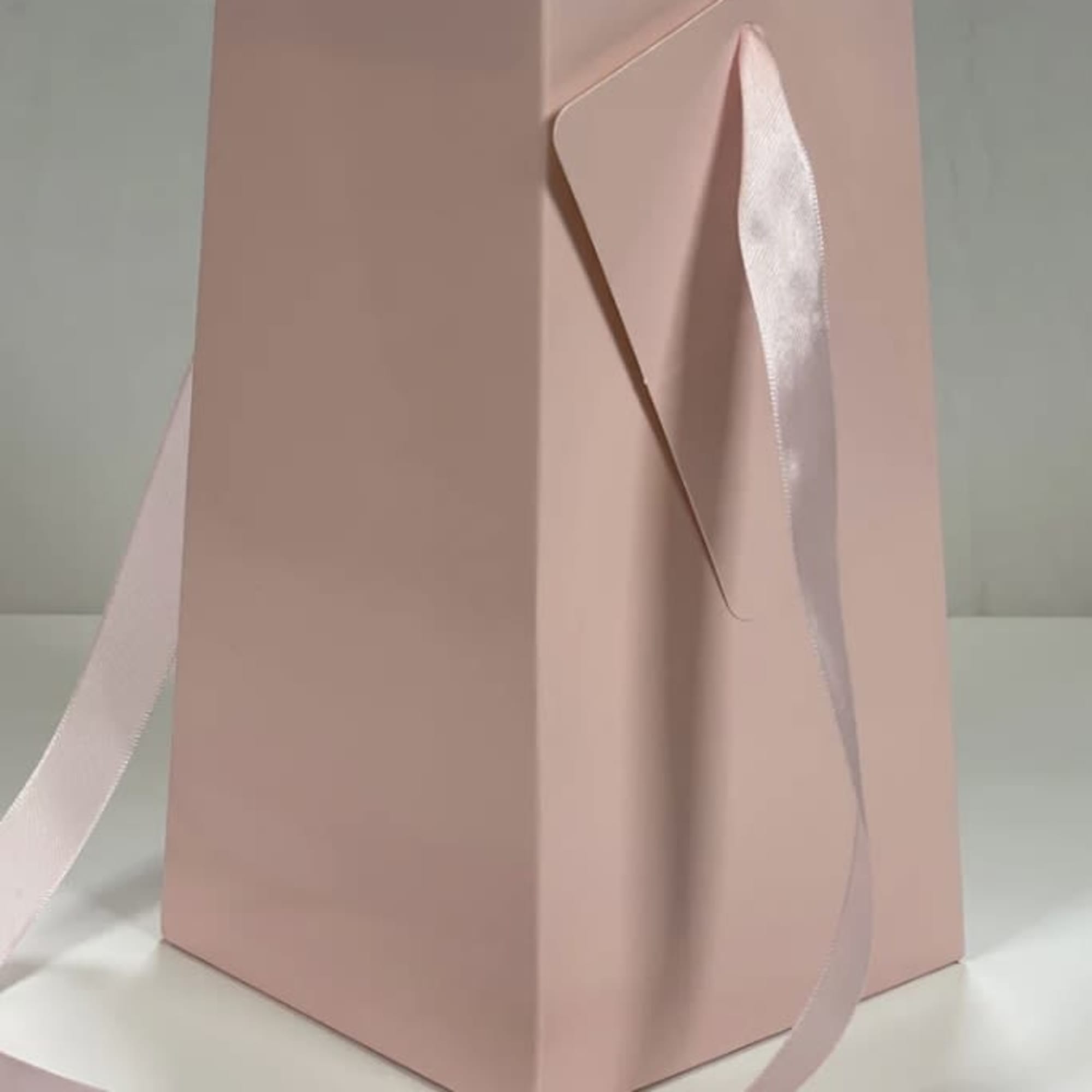 Tall blush pink gift bag or box with a long matching satin ribbon handle on a white surface