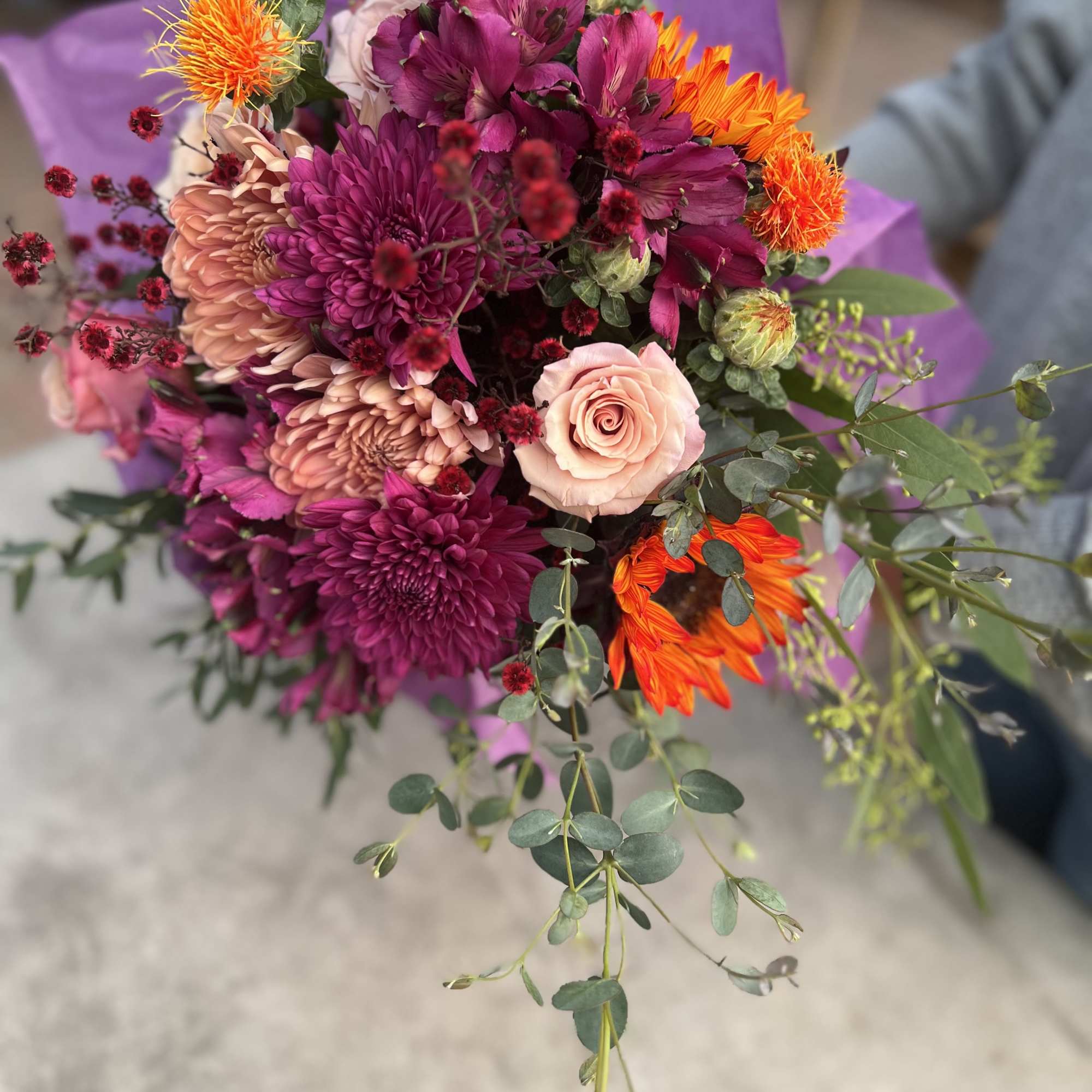 Hand-tied bouquet of pink roses, purple mums, and orange flowers wrapped in purple paper