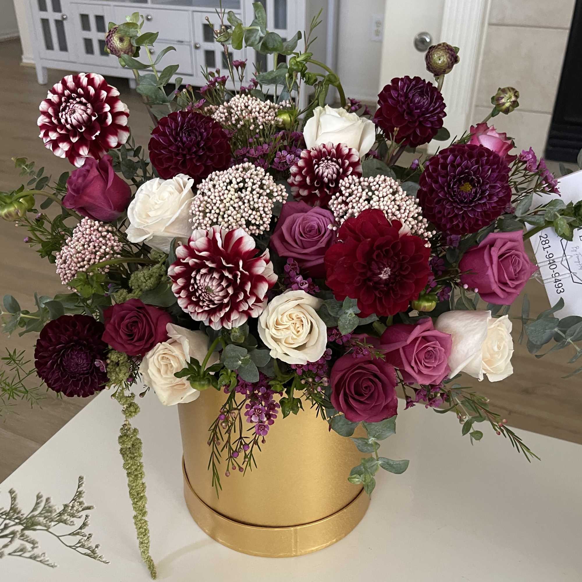 Burgundy dahlias and pink and cream roses arranged in a gold cylinder box.