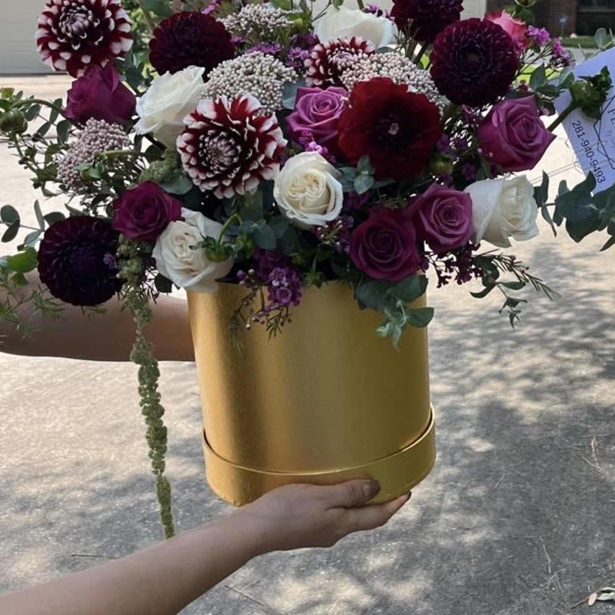 Arrangement of burgundy dahlias and purple and white roses in a round gold hatbox container