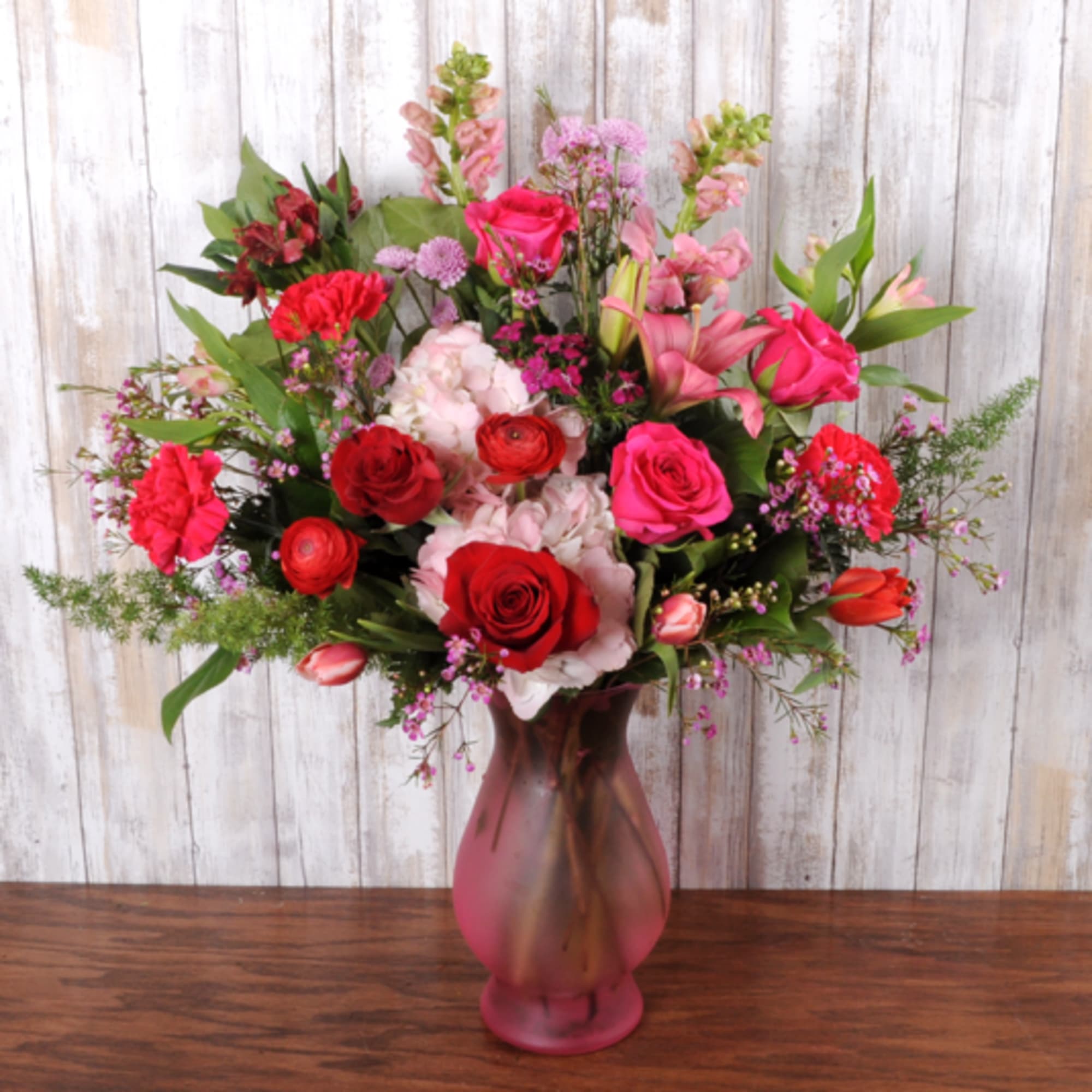 Tall arrangement of red and hot pink roses, carnations, lilies and tulips in a frosted pink glass vase