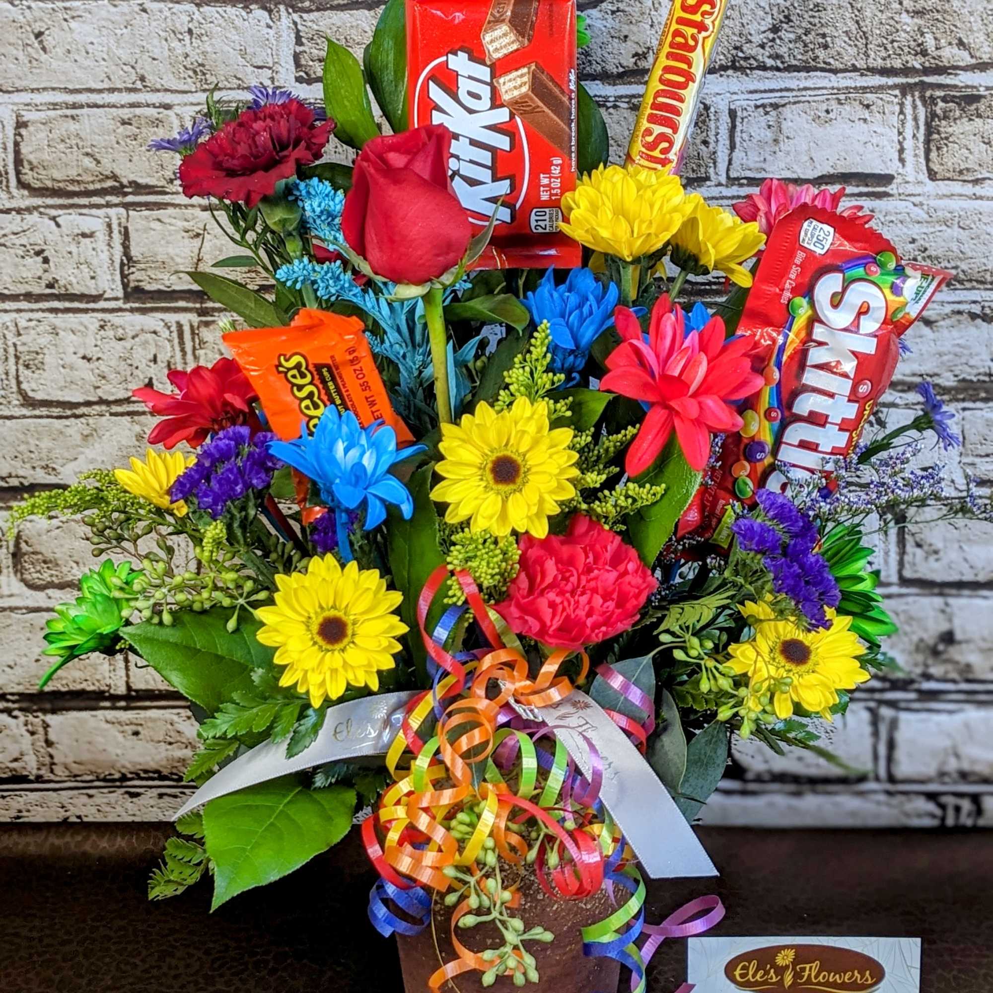 Colorful candy and flower arrangement in a glass vase with roses, carnations, and bright daisies.