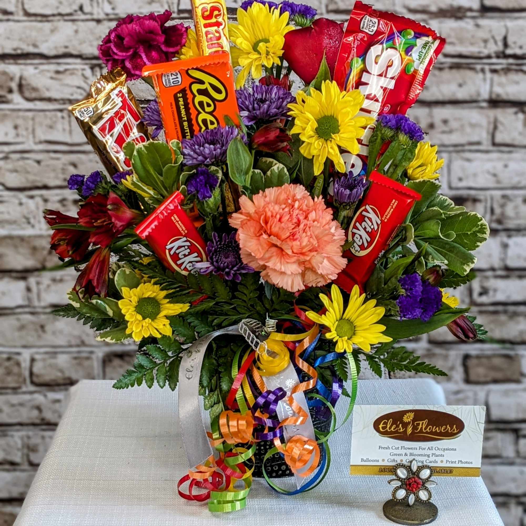 Mixed bouquet of bright flowers arranged in a mug with assorted candy bars and curling ribbons.