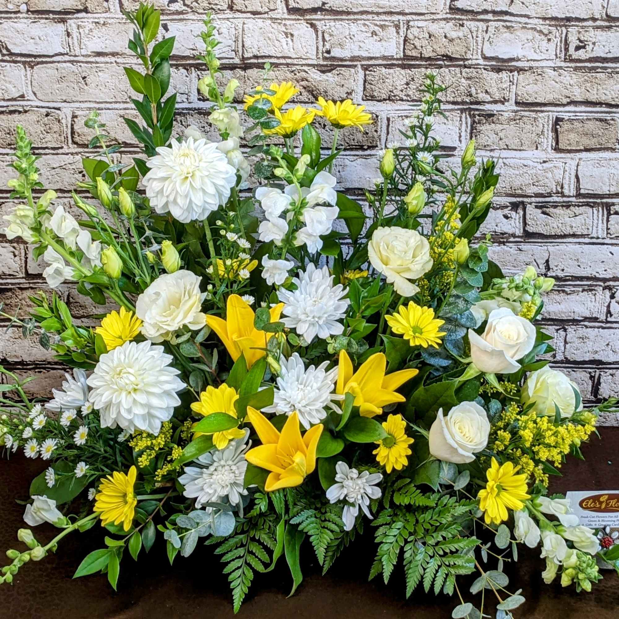 Tall fan-shaped arrangement of white roses, mums, and snapdragons with yellow lilies and daisies.