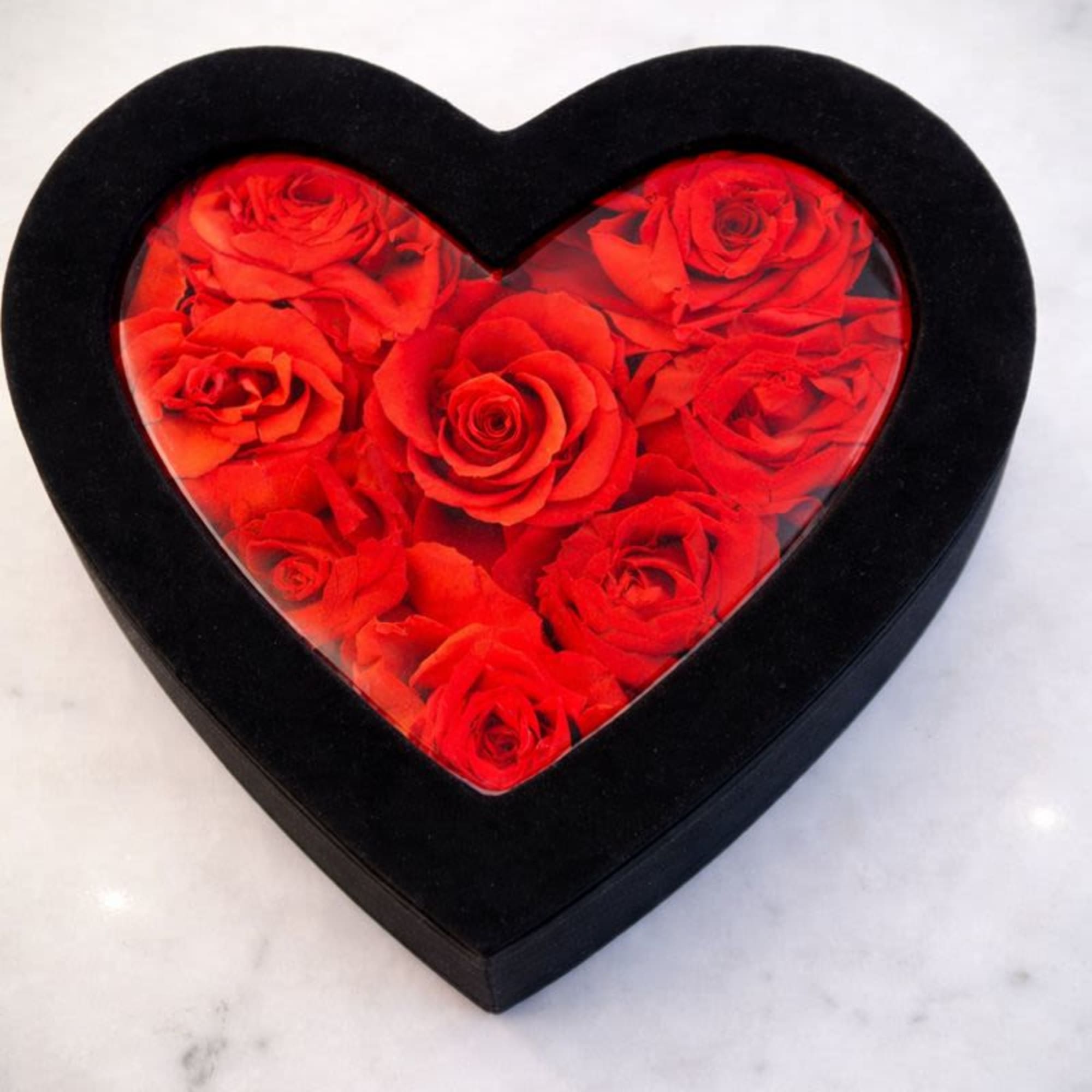 Heart-shaped black box filled with red roses under a clear lid on a light surface