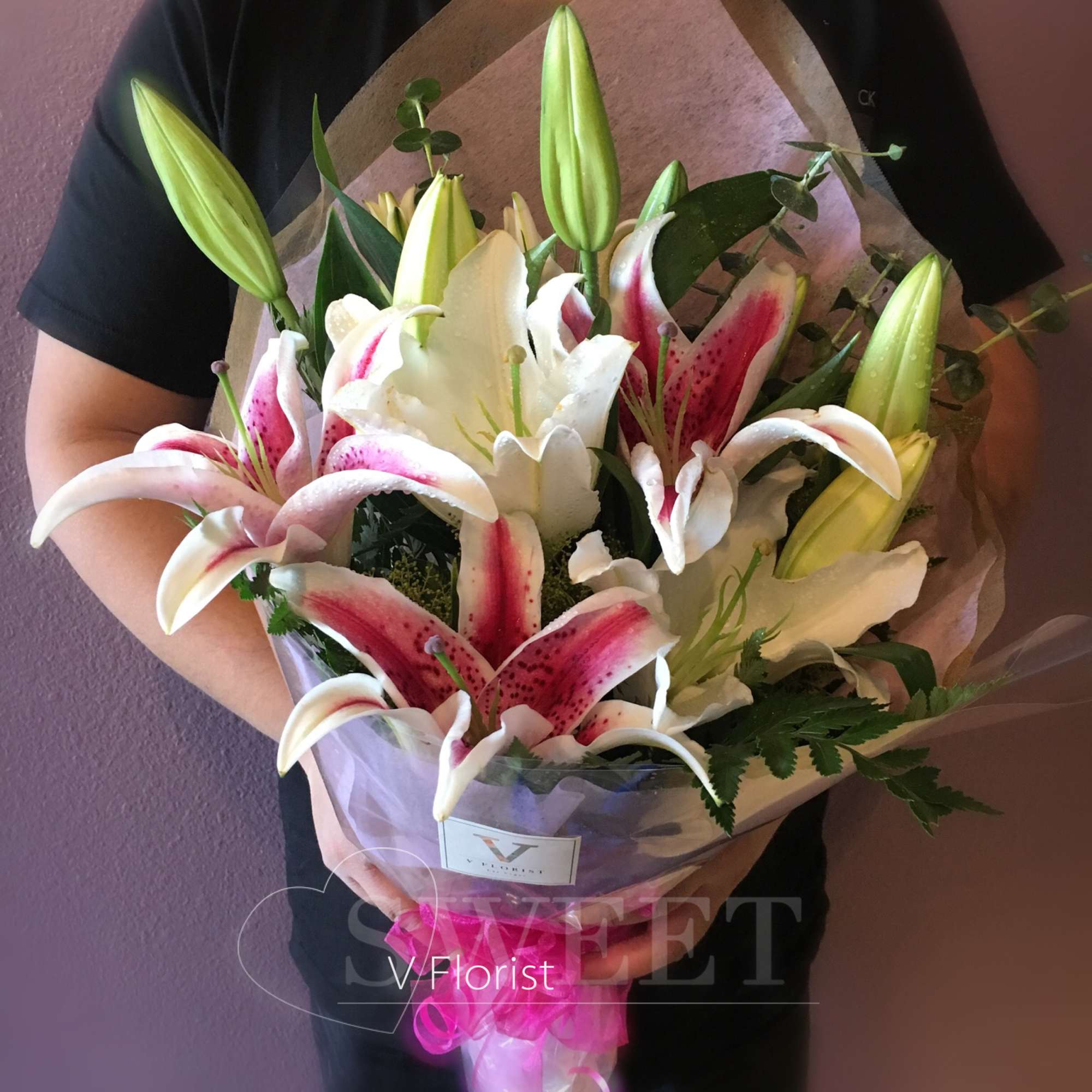 Bouquet of white and pink lilies with green buds wrapped in tissue and ribbon, held in front of a mauve wall