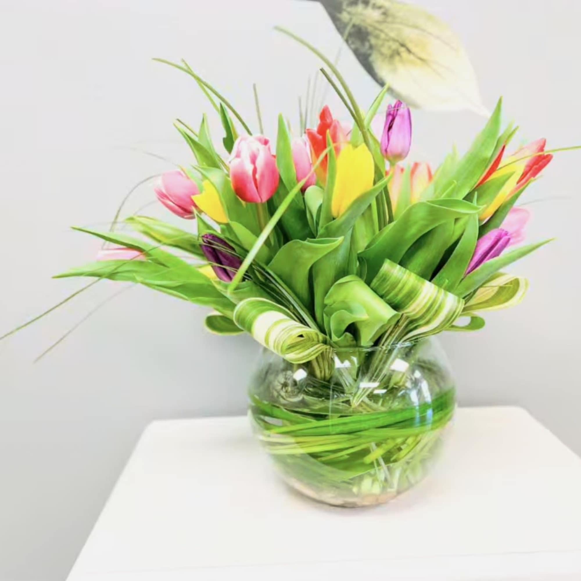 Round glass vase filled with multicolored tulips and decorative looped greenery.