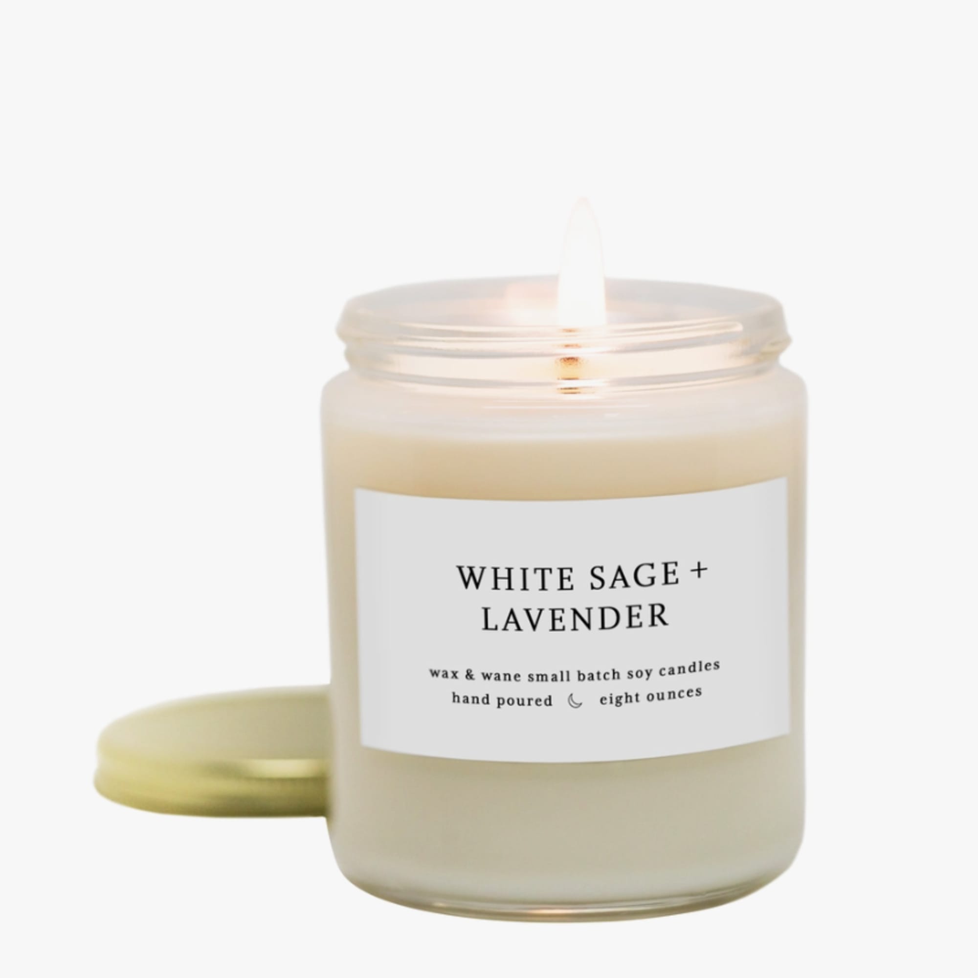 White jar candle labeled White Sage + Lavender with lit flame and gold lid beside it