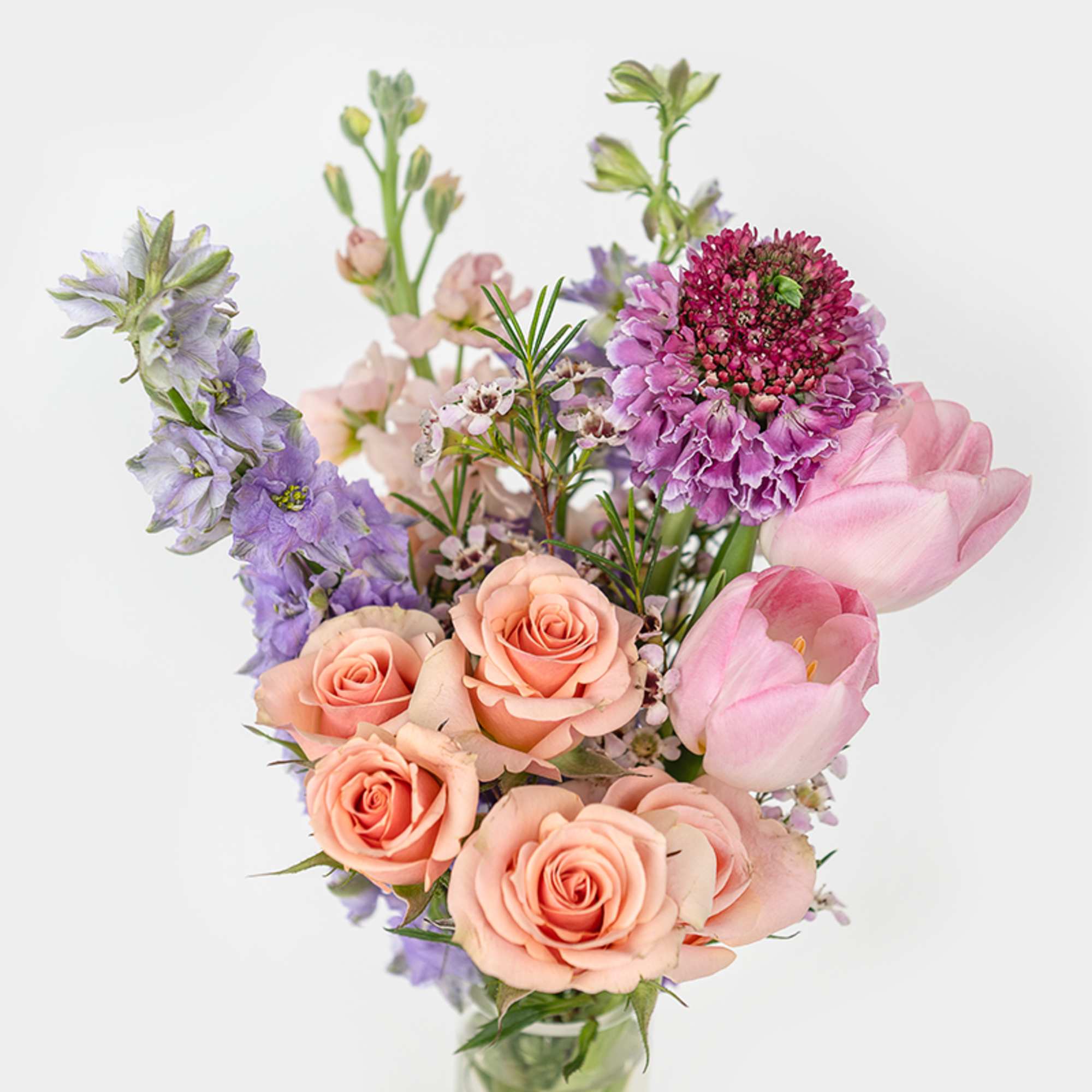 Pastel bouquet of peach roses, pink tulips, and purple blooms arranged in a clear glass vase