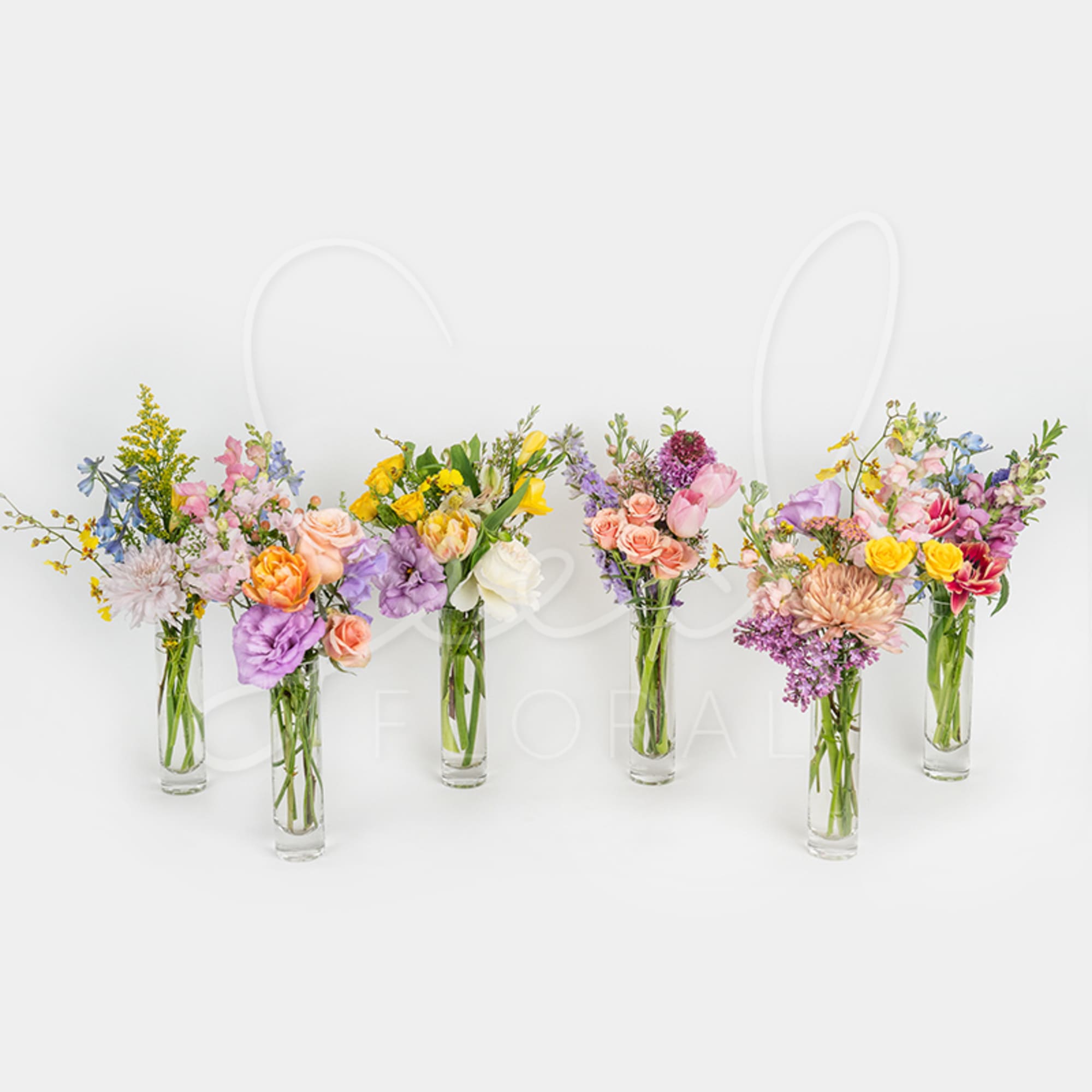 Six slim glass vases arranged in a row with assorted pastel and bright mixed flowers.