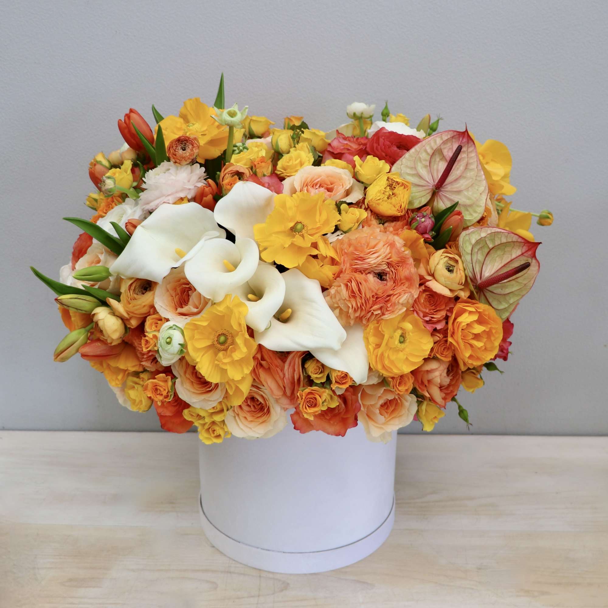 Lush arrangement of white calla lilies with orange and yellow roses and ranunculus in a round white hatbox container