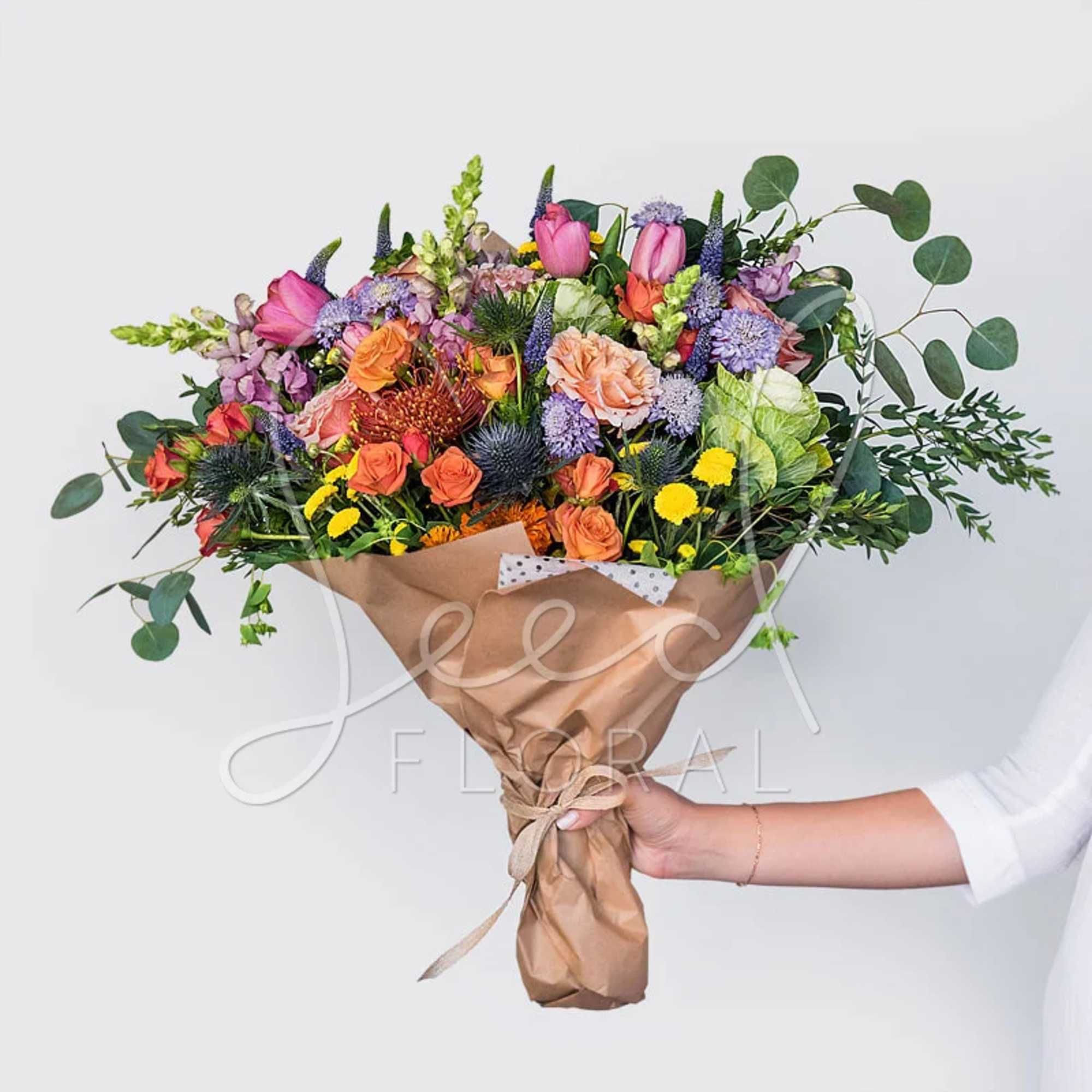 Large hand-tied bouquet of mixed bright flowers wrapped in brown kraft paper and held in one hand.