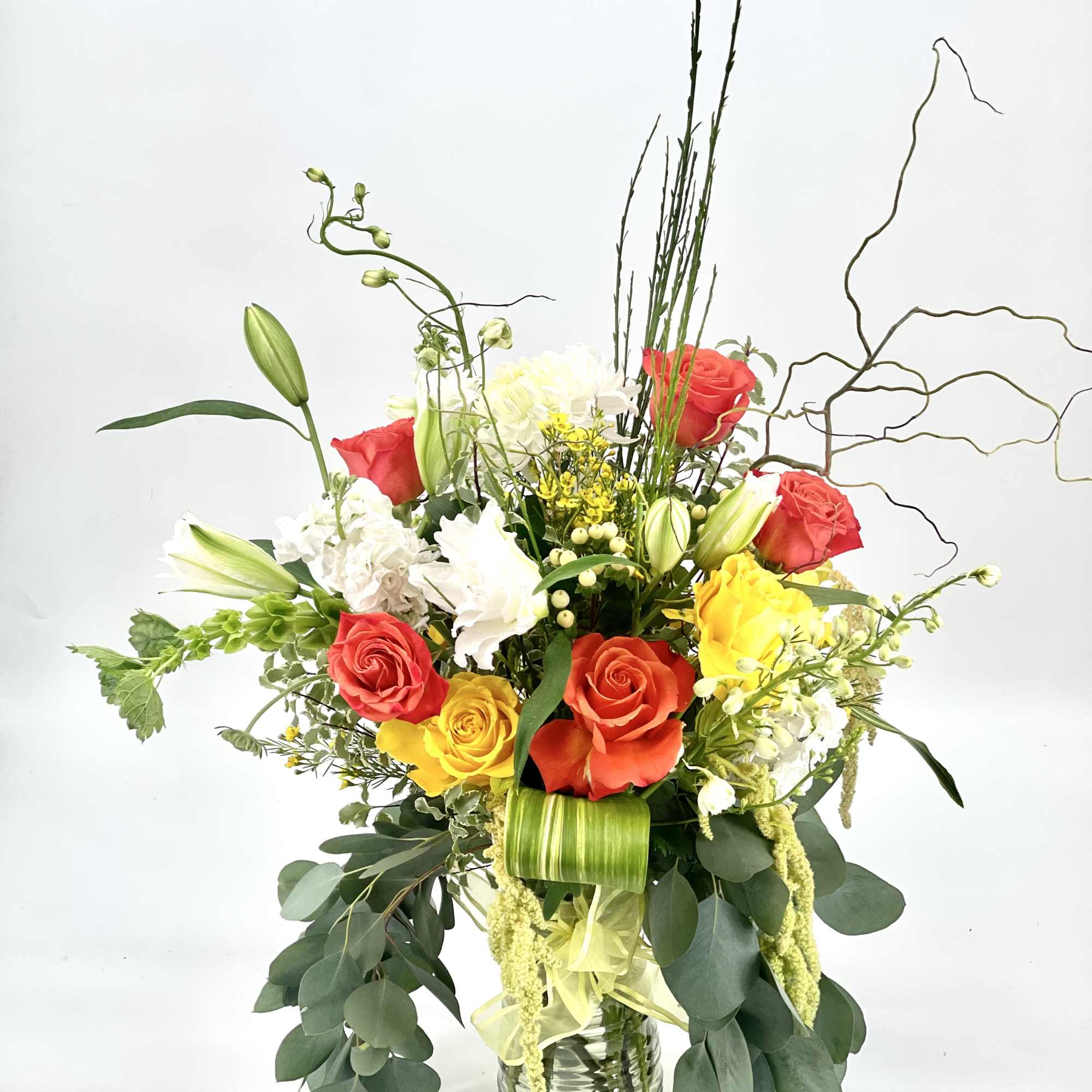 Tall mixed arrangement with orange and yellow roses, white lilies, and white blooms in a clear glass vase