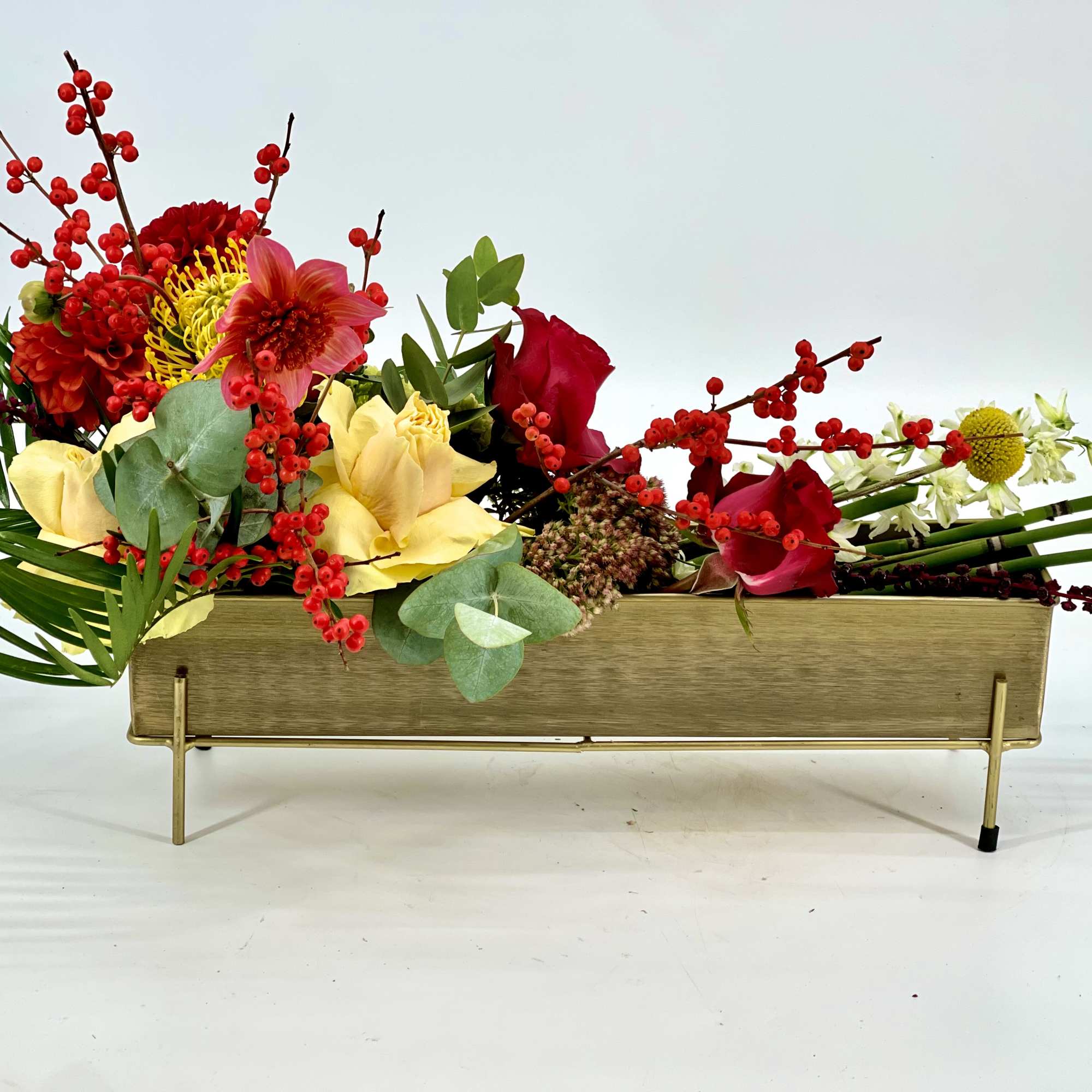 Low rectangular gold centerpiece with red, yellow, and white flowers and berries in an asymmetrical design.