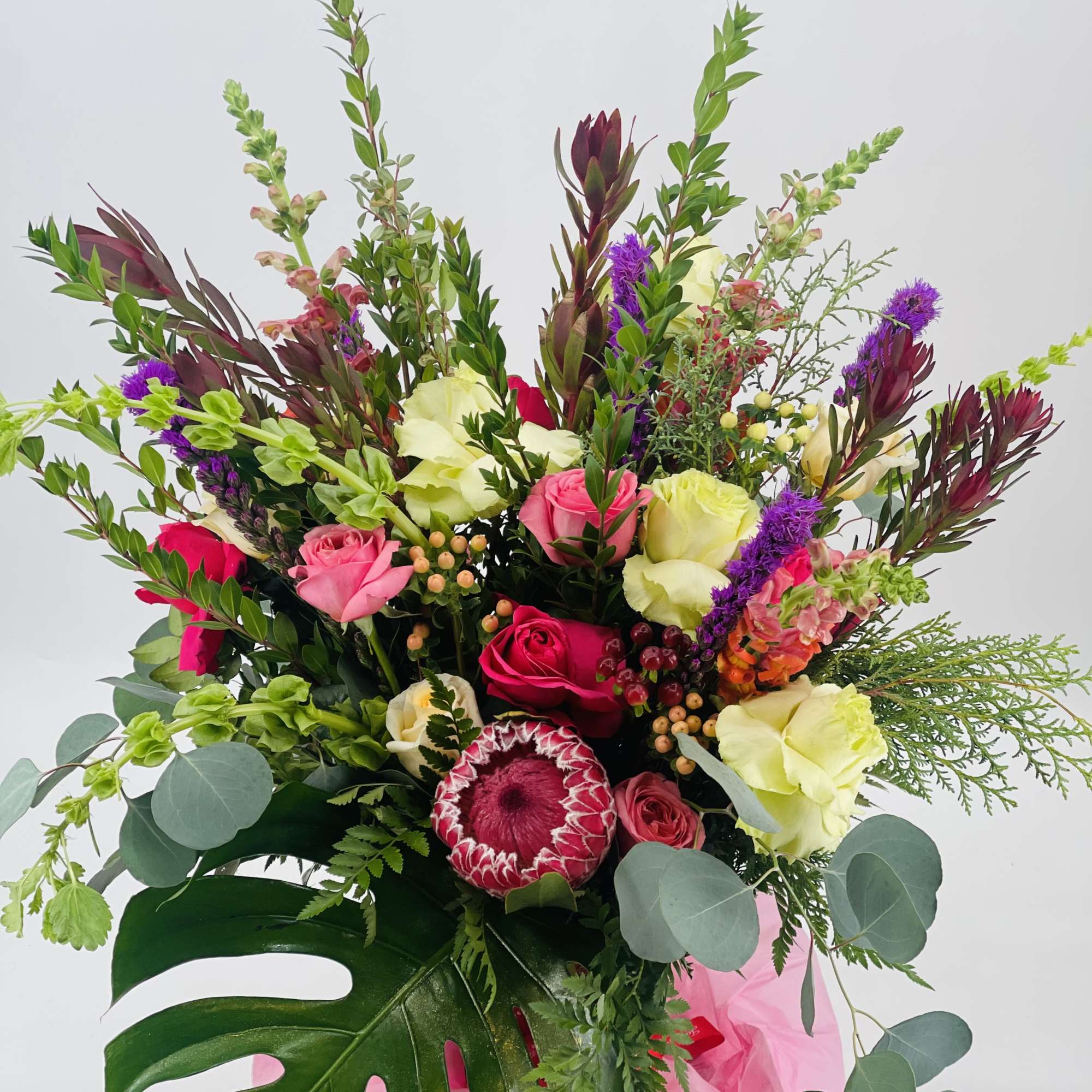 Tall mixed arrangement with pink roses, protea, cream blooms and purple accents in a white box with a large monstera leaf.