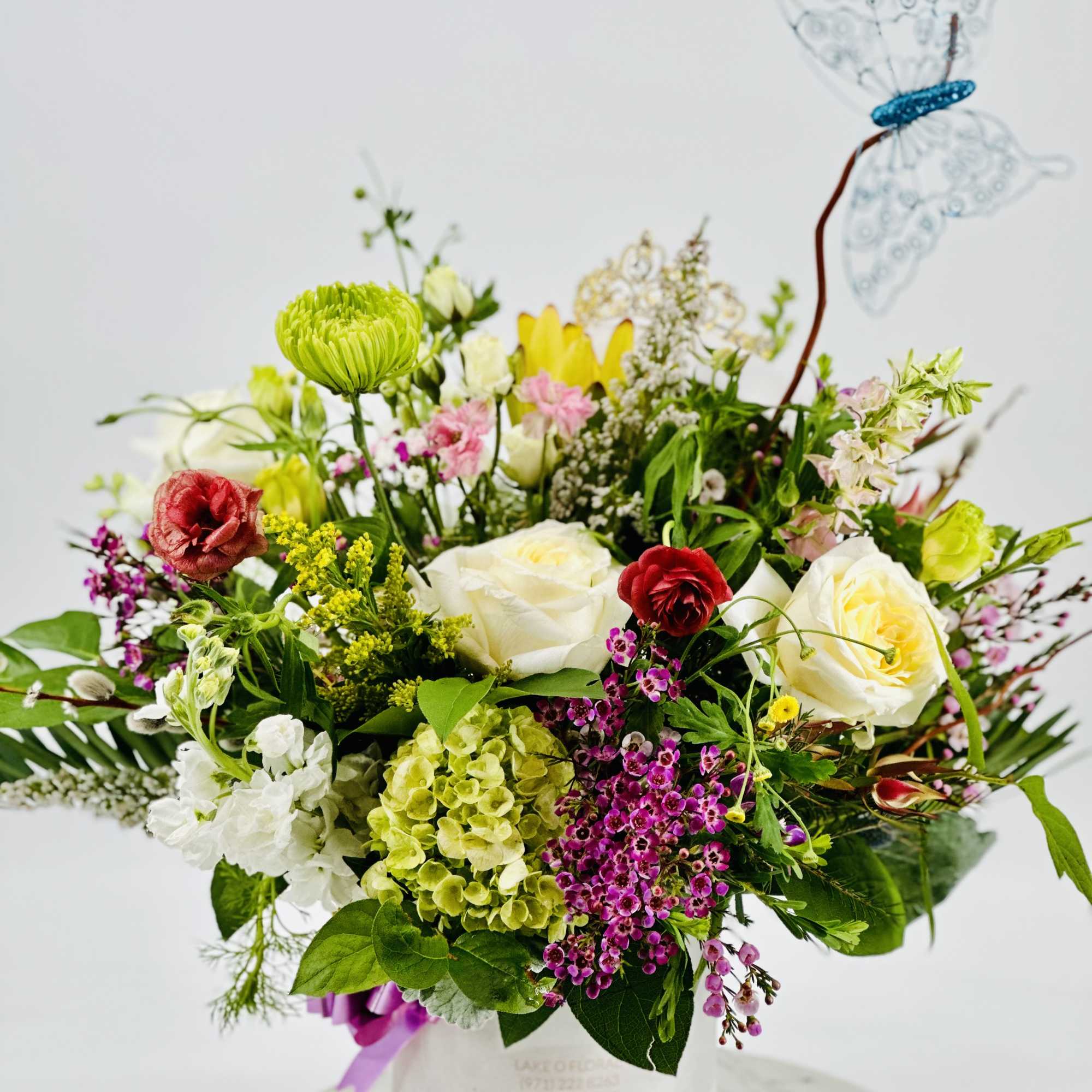Colorful mixed flower arrangement with roses, hydrangea, and a blue decorative butterfly in a white vase.