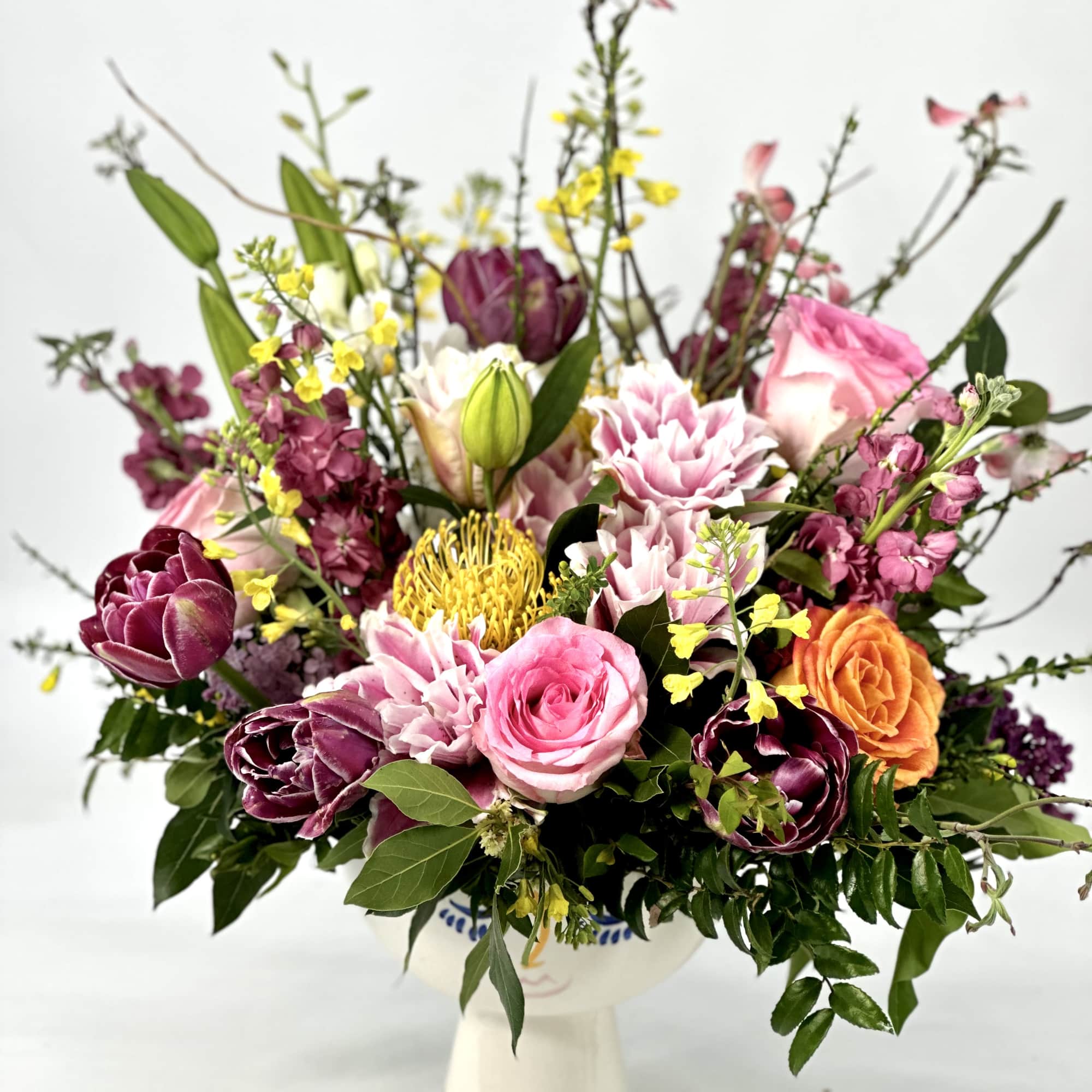 Lush mixed arrangement of pink roses, purple tulips, dahlias and protea in a white face vase.