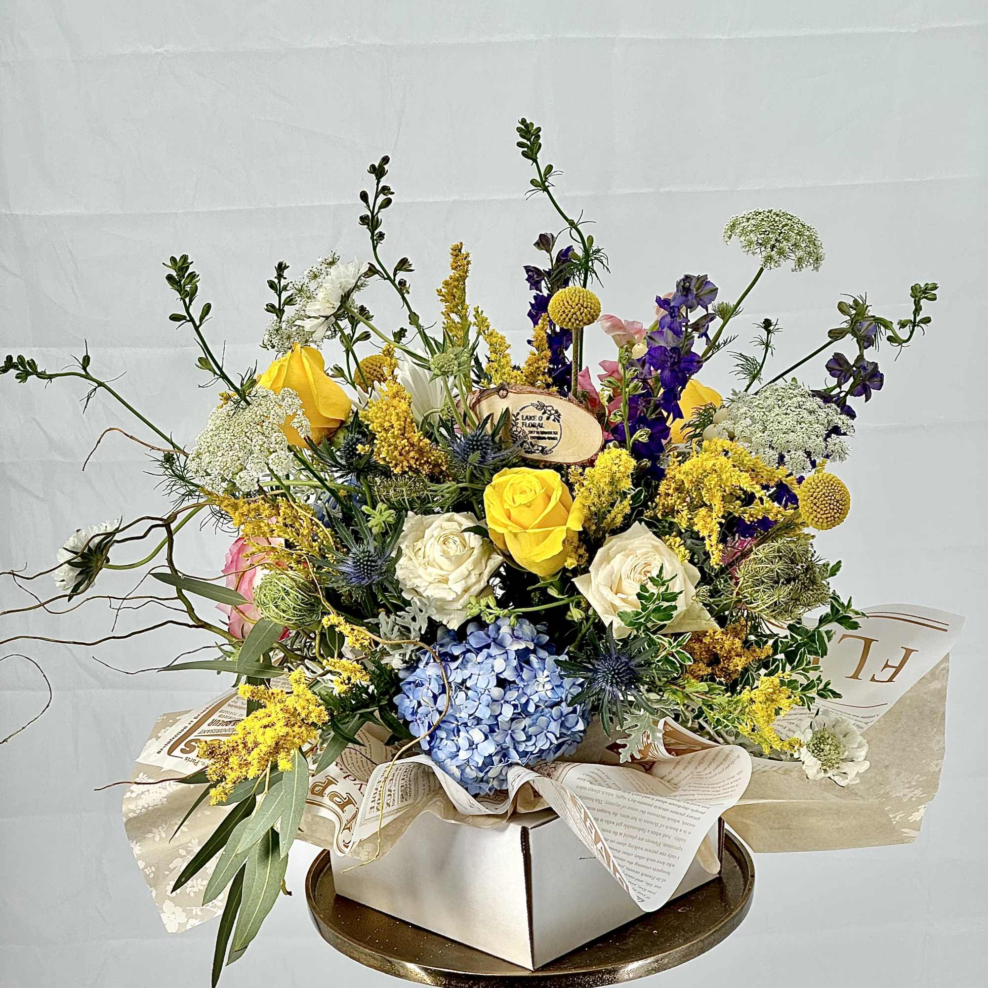 Colorful arrangement of yellow roses, blue hydrangea, and mixed blooms in a paper-wrapped box.