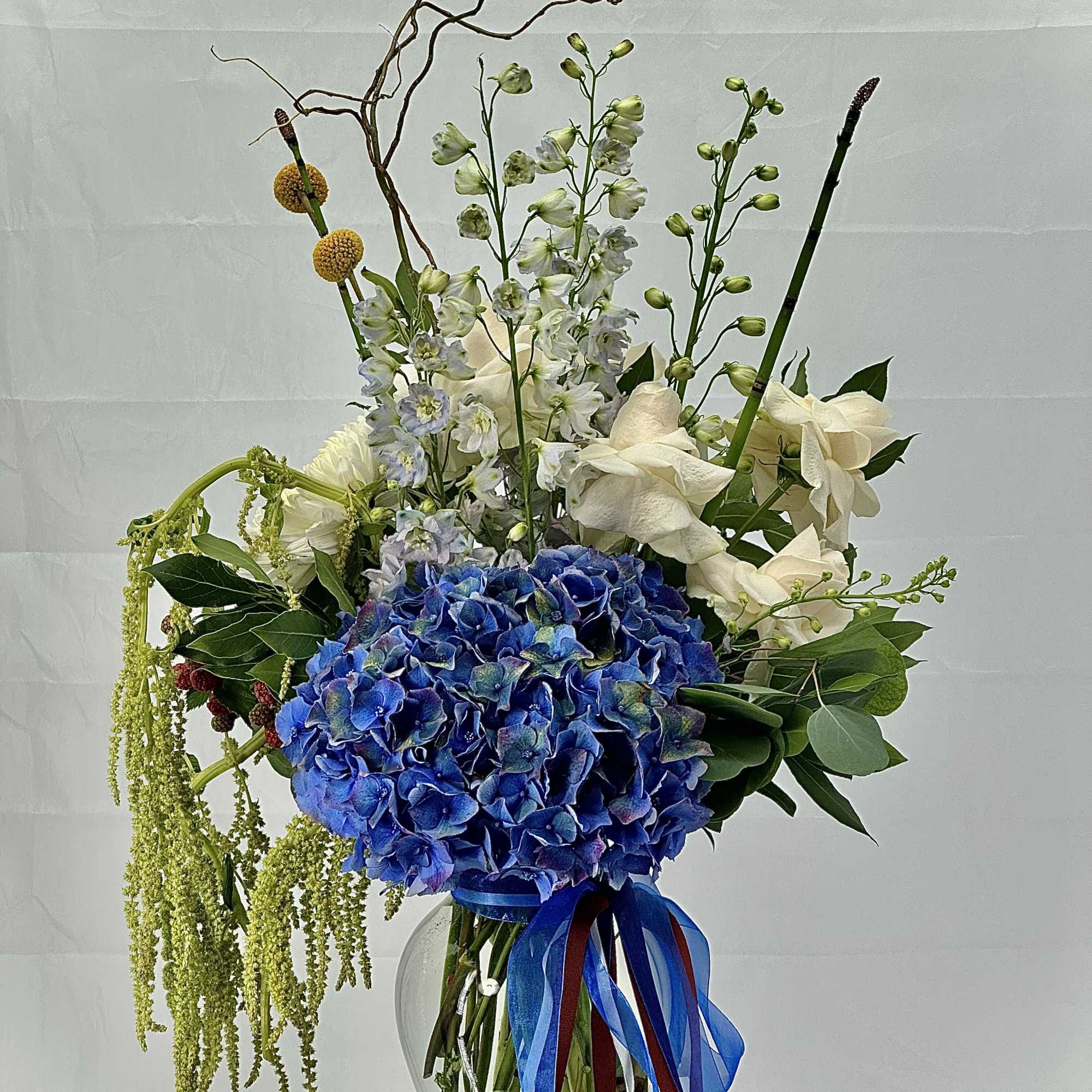 Tall arrangement with blue hydrangeas and white flowers in a glass vase with ribbons