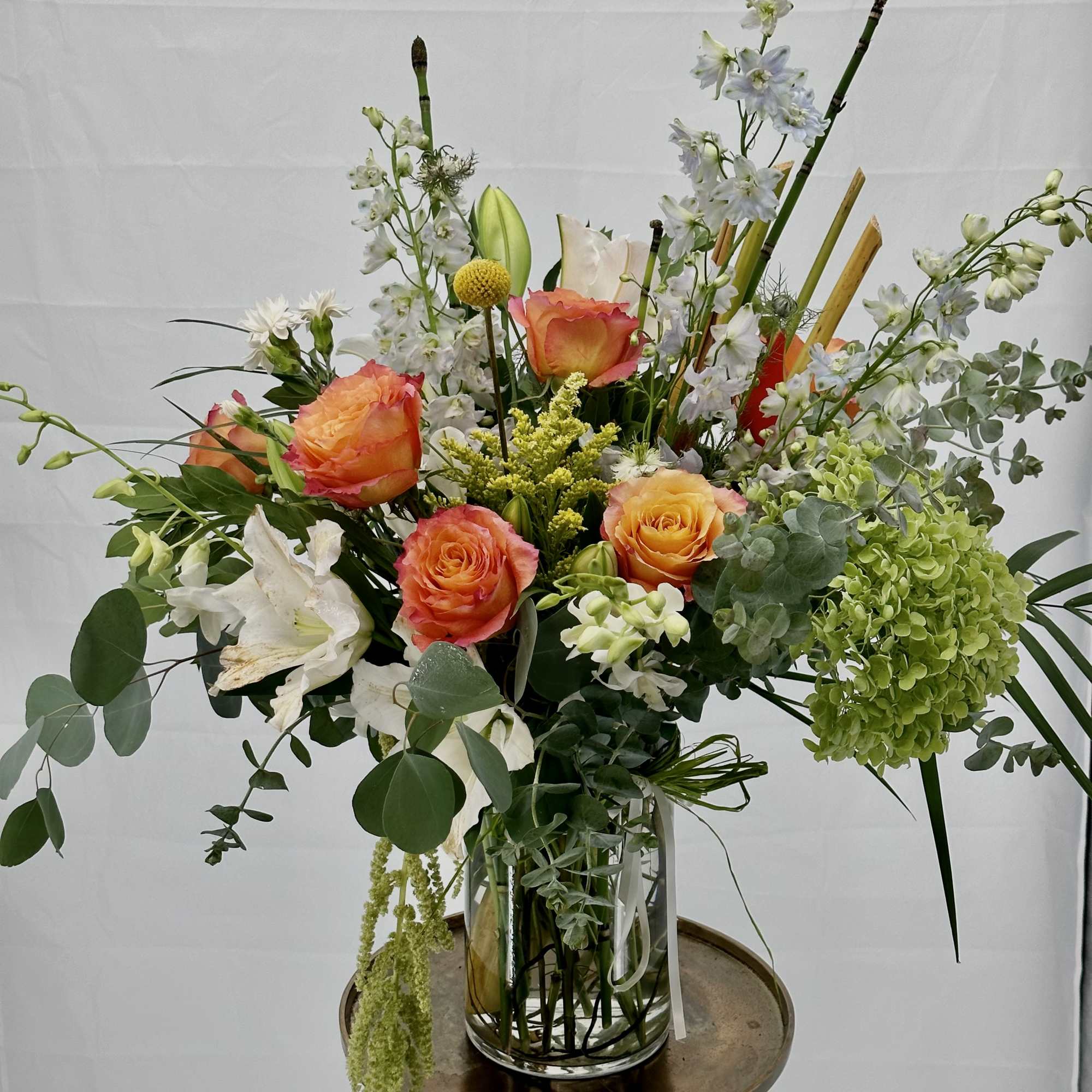 Tall mixed arrangement of orange roses, white lilies, and green hydrangea in a clear glass vase