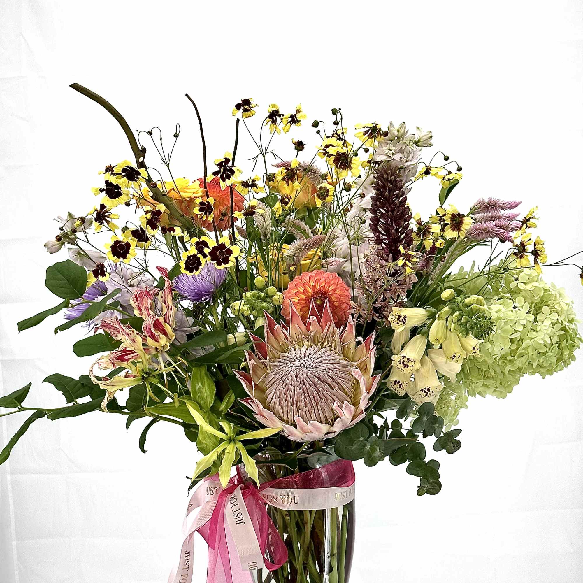 Tall mixed bouquet with protea, hydrangea, dahlias and wildflowers in a clear glass vase with pink ribbons.