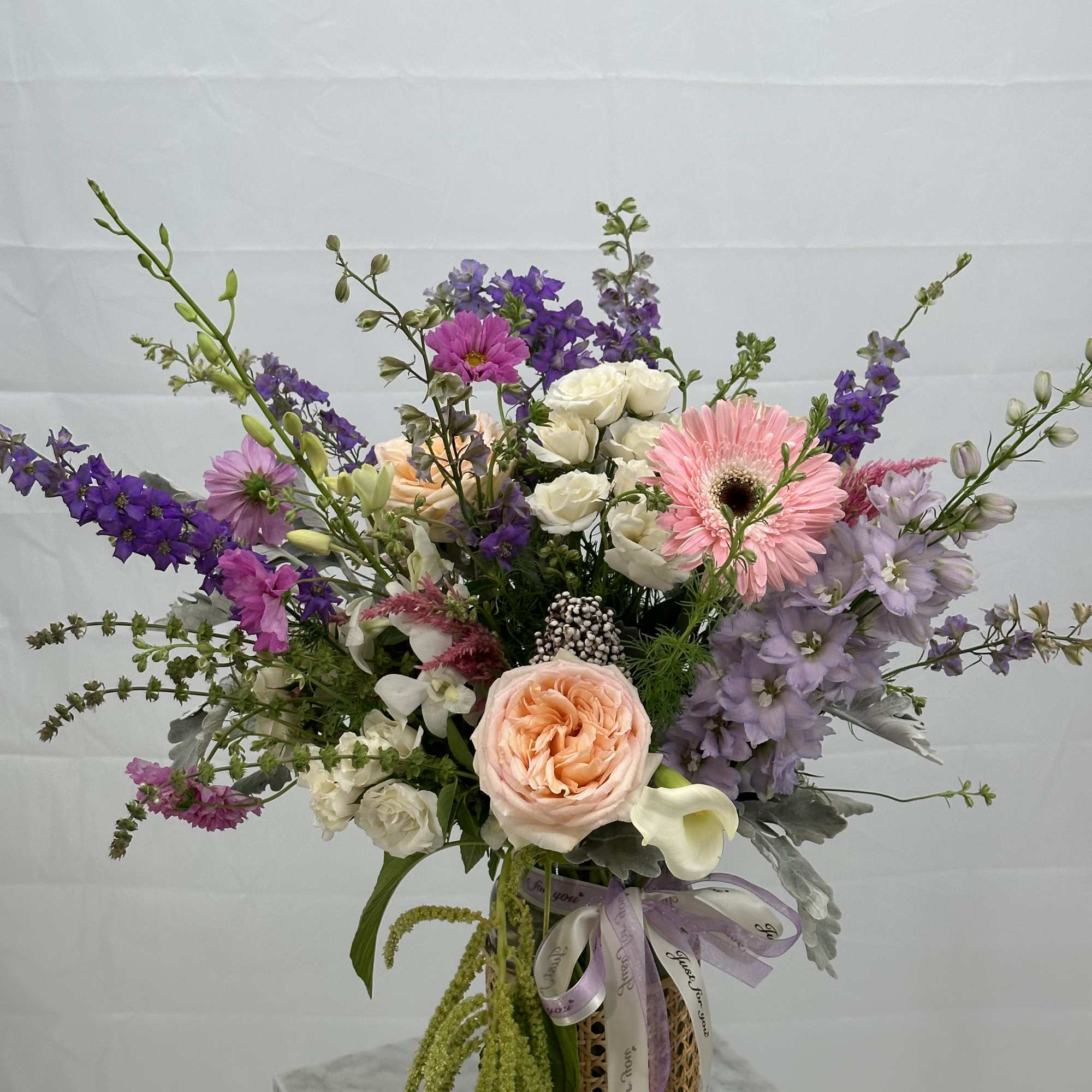 Tall garden-style arrangement of peach, pink, and purple flowers in a glass vase with ribbon
