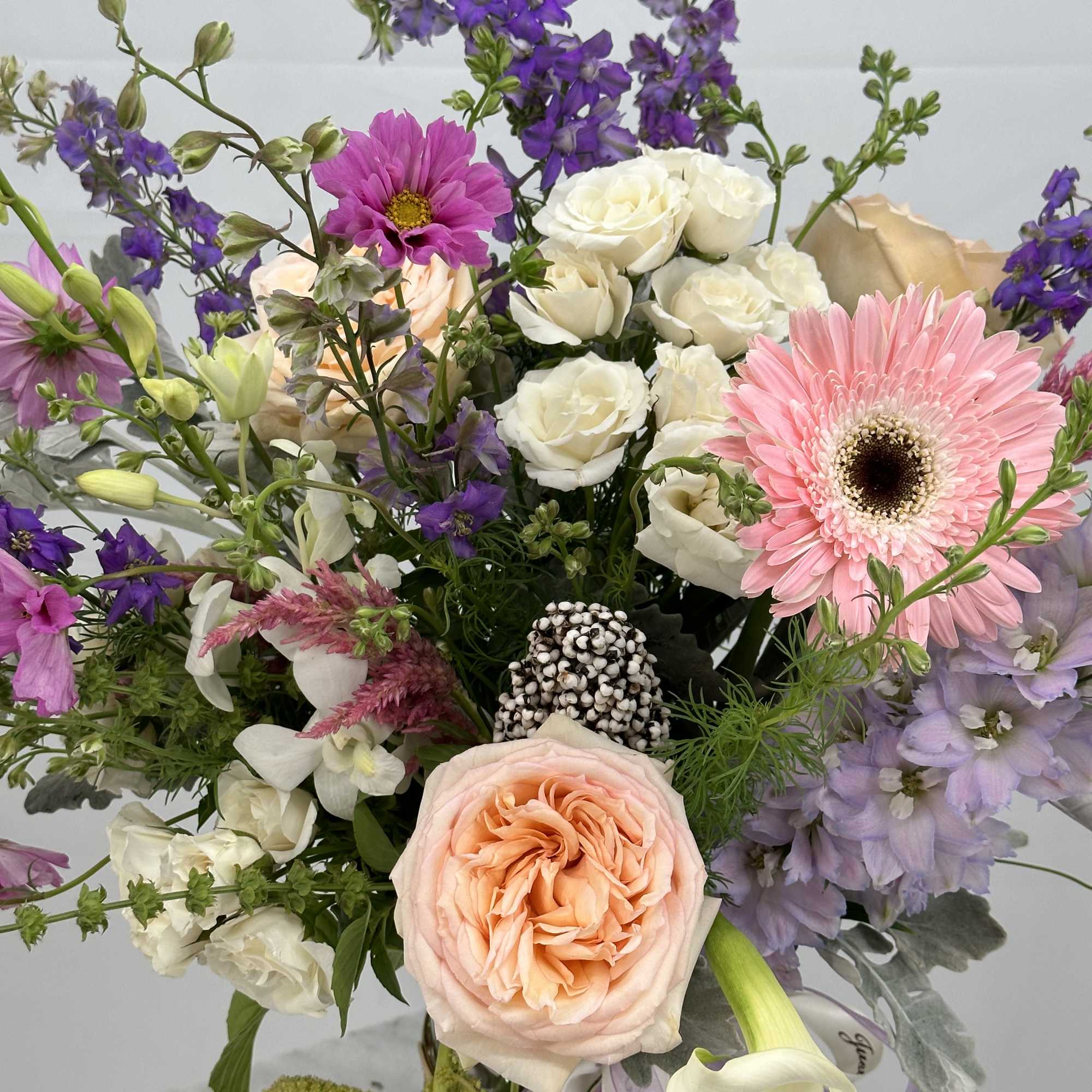 Loose garden-style bouquet of peach roses, pink gerbera, and purple flowers in a woven basket with ribbons
