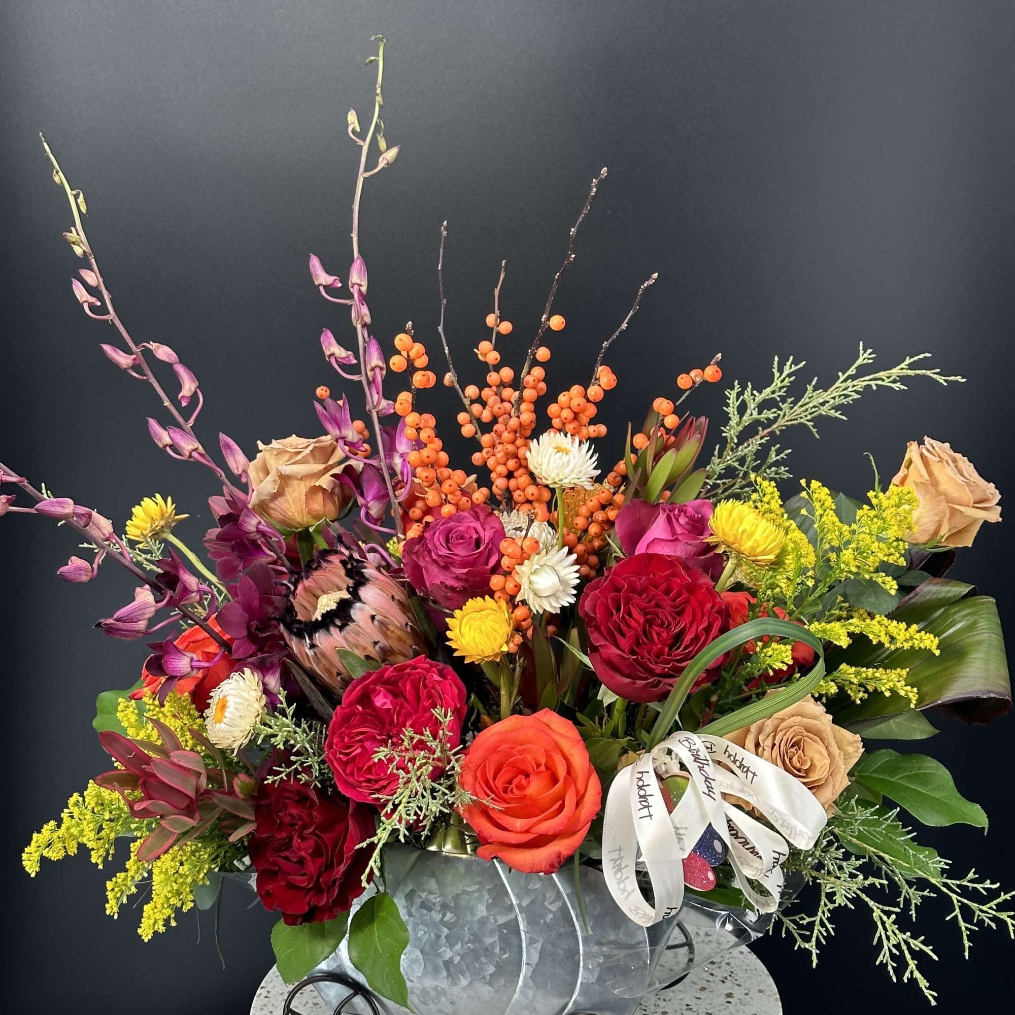 Bright mixed fall flower arrangement with roses, protea, and berries in a metal wagon container.