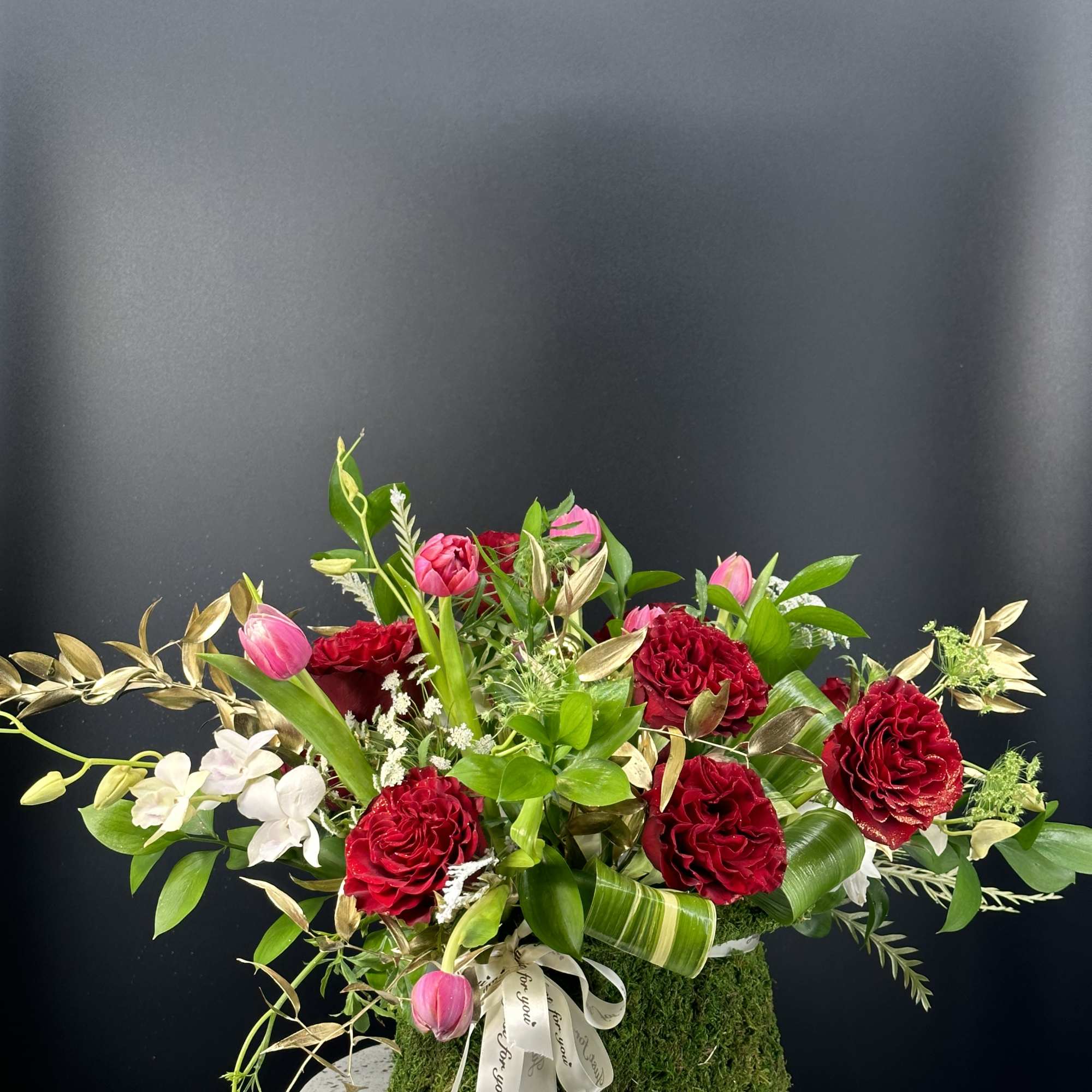 Low arrangement of red roses, pink tulips, and white orchids in a moss-covered container with ribbon.