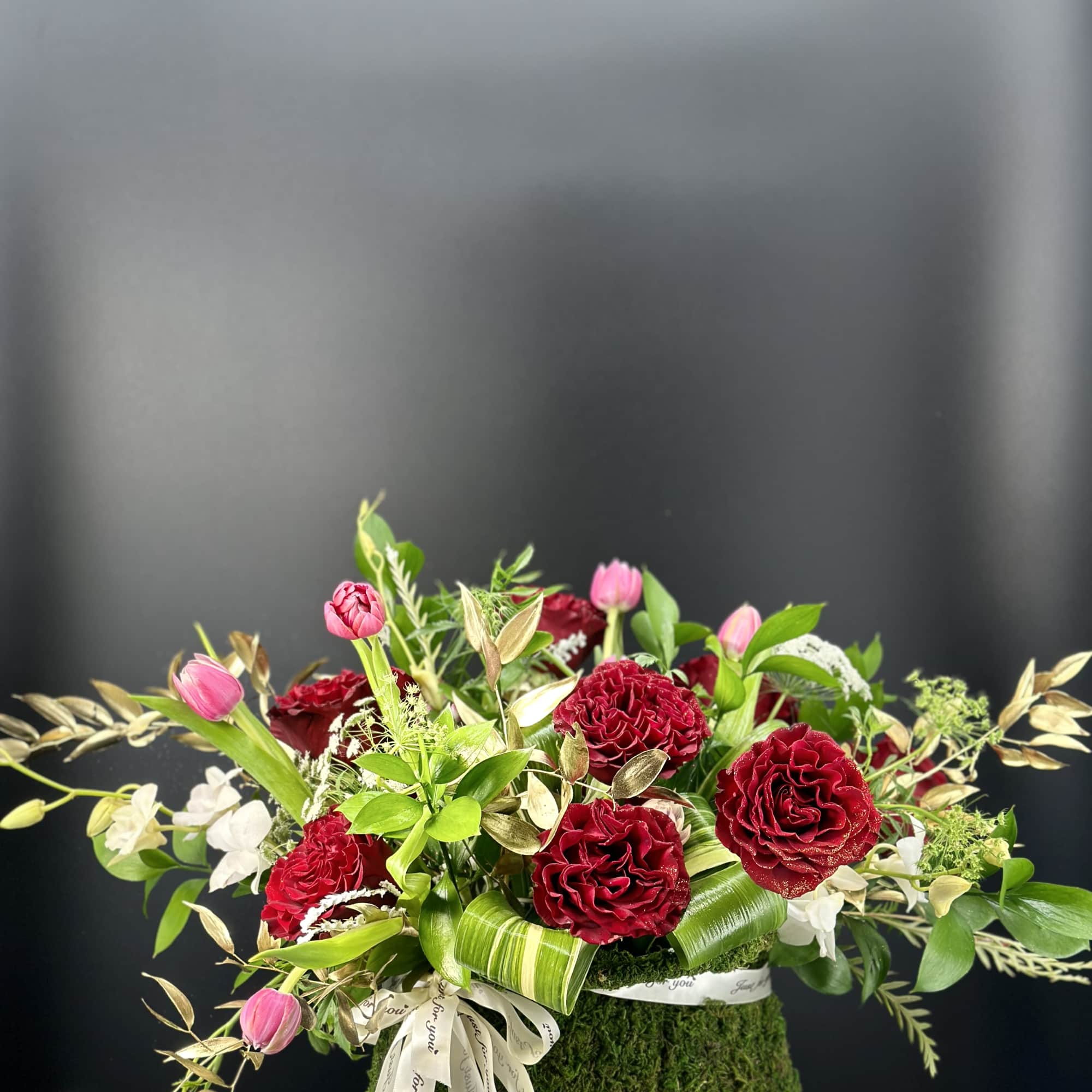 Low arrangement of red roses and pink tulips in a moss-covered container with ribbon bow