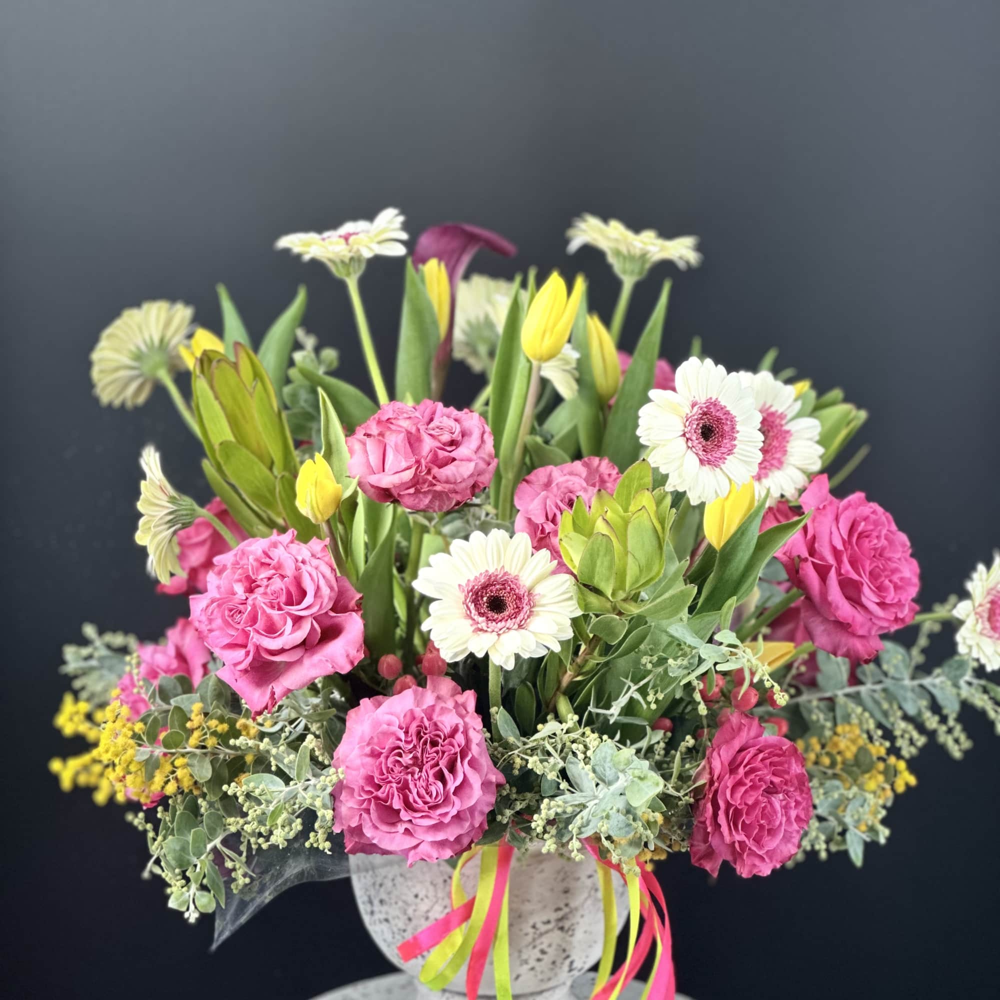 Bright arrangement of pink carnations, white gerbera daisies and yellow tulips in a stone urn with colorful ribbons