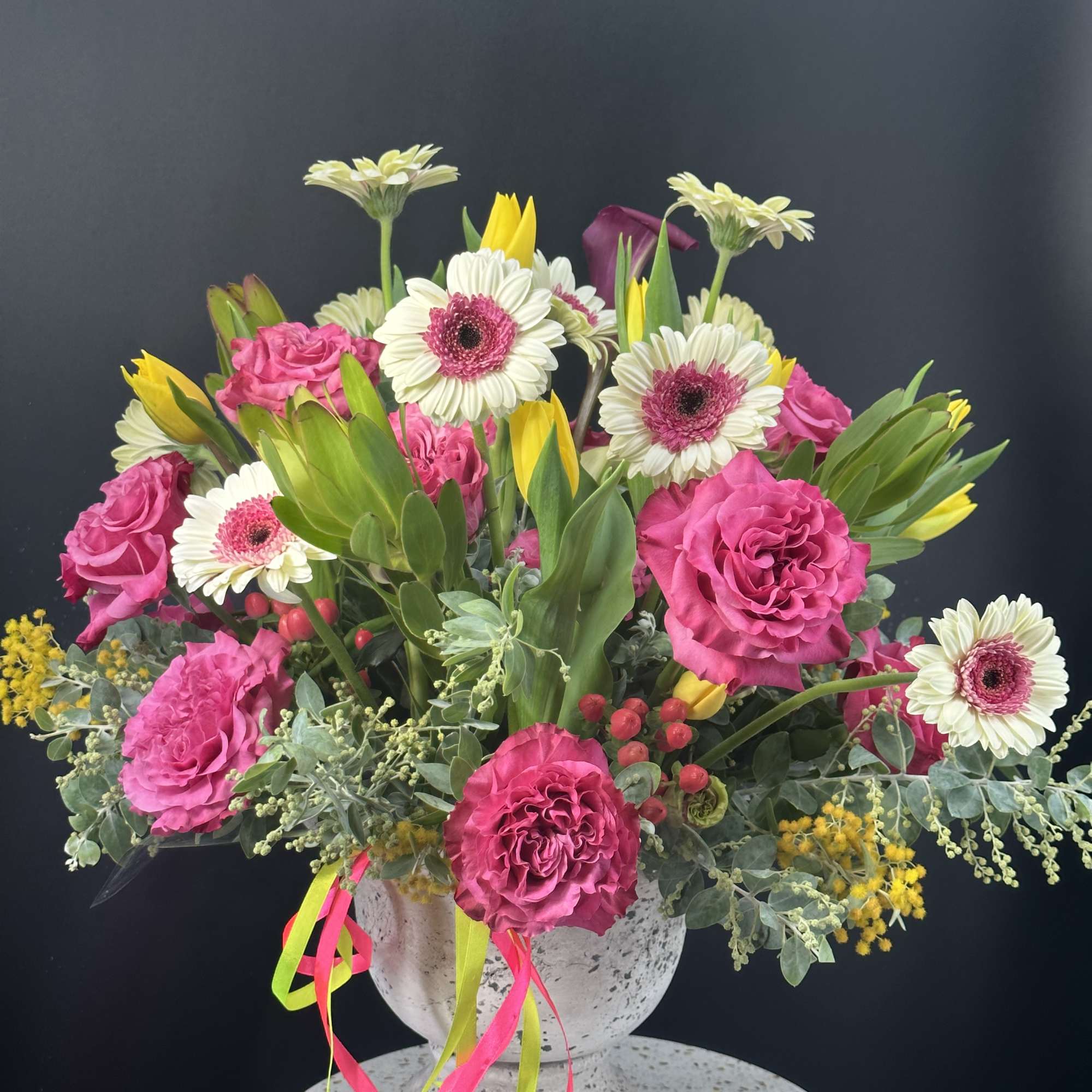 Bright arrangement of hot pink carnations, white gerbera daisies and yellow tulips in a stone urn with colorful ribbons.