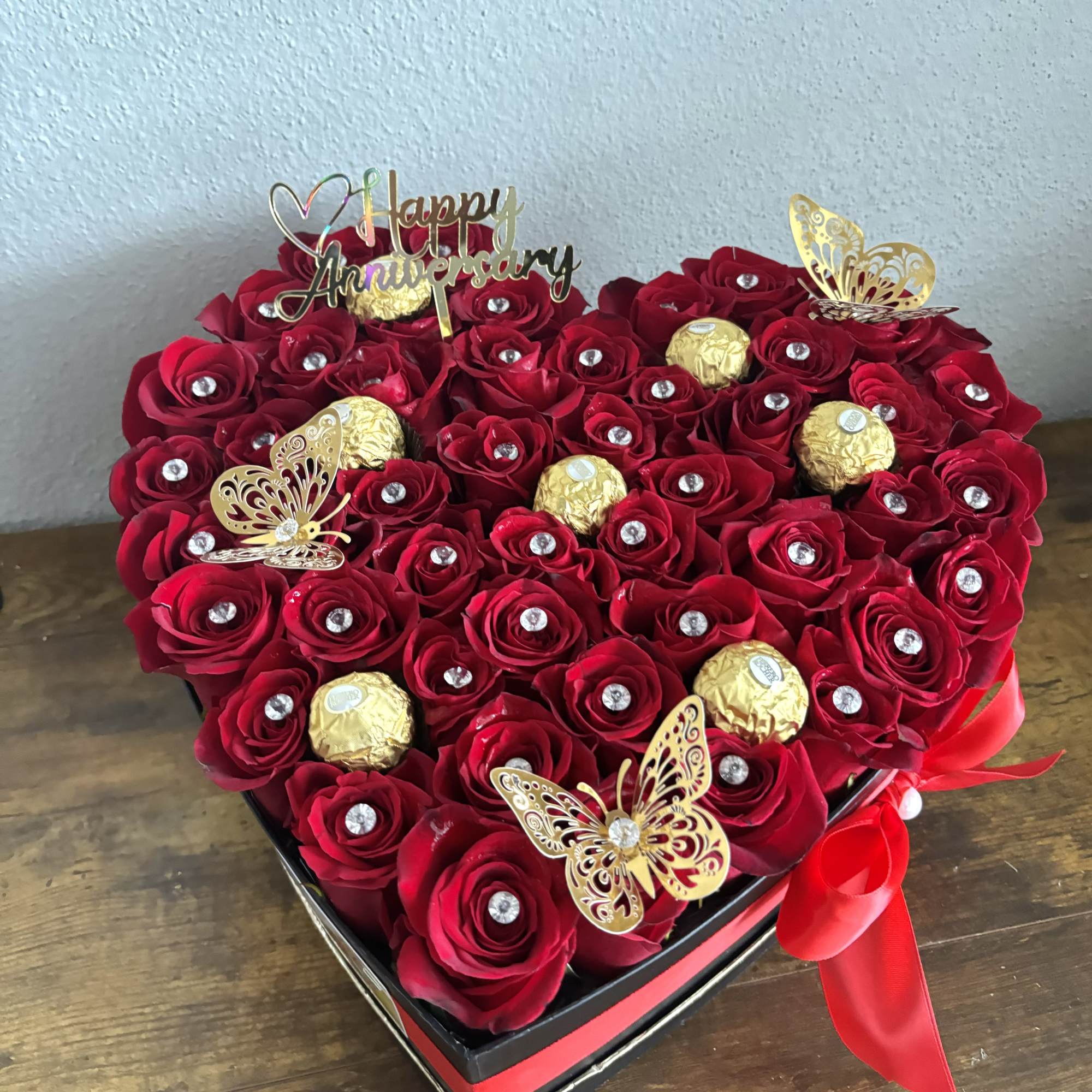 Heart-shaped box of red roses with chocolates and a Happy Anniversary topper