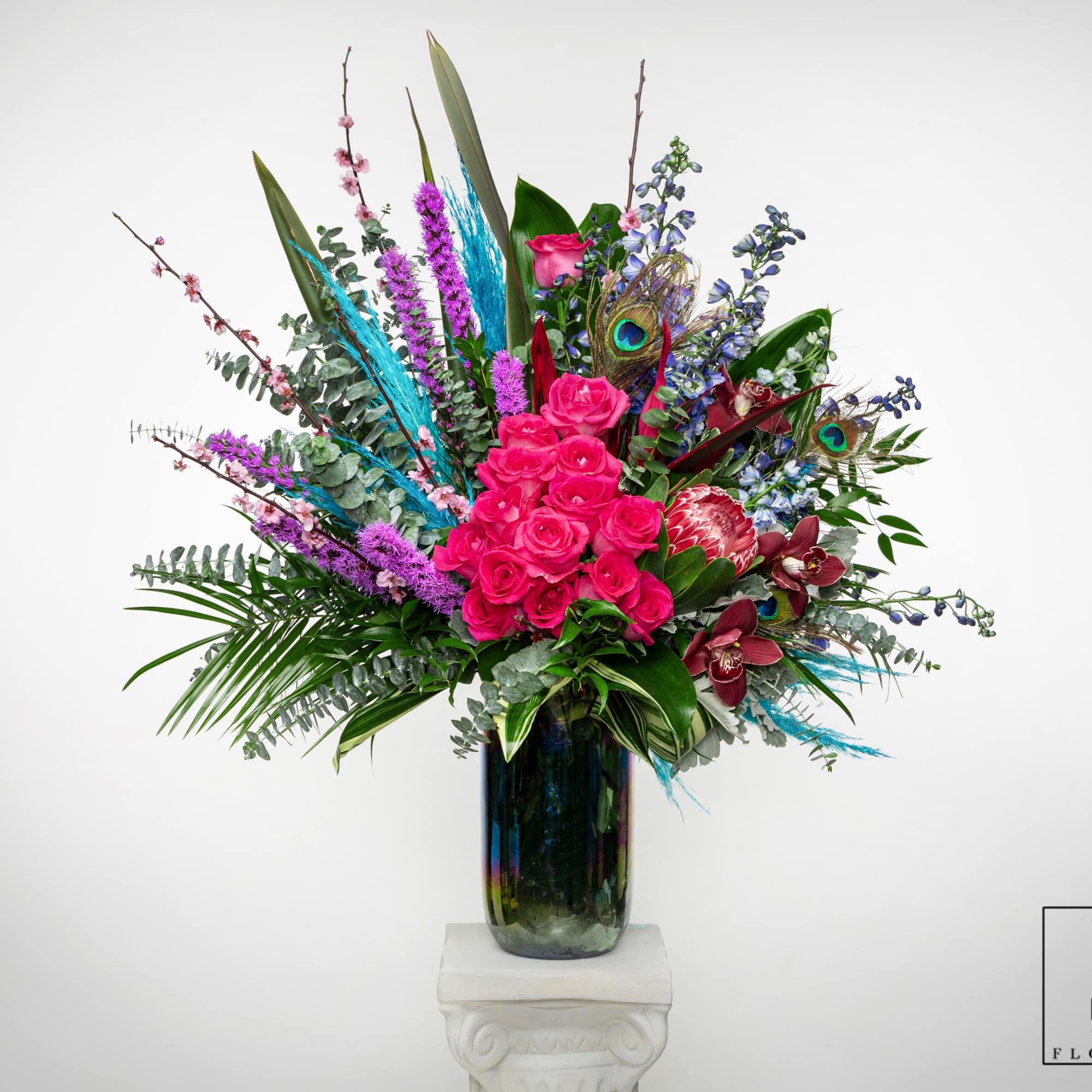 Tall arrangement of hot pink roses, orchids, protea, and peacock feathers in a dark glass vase