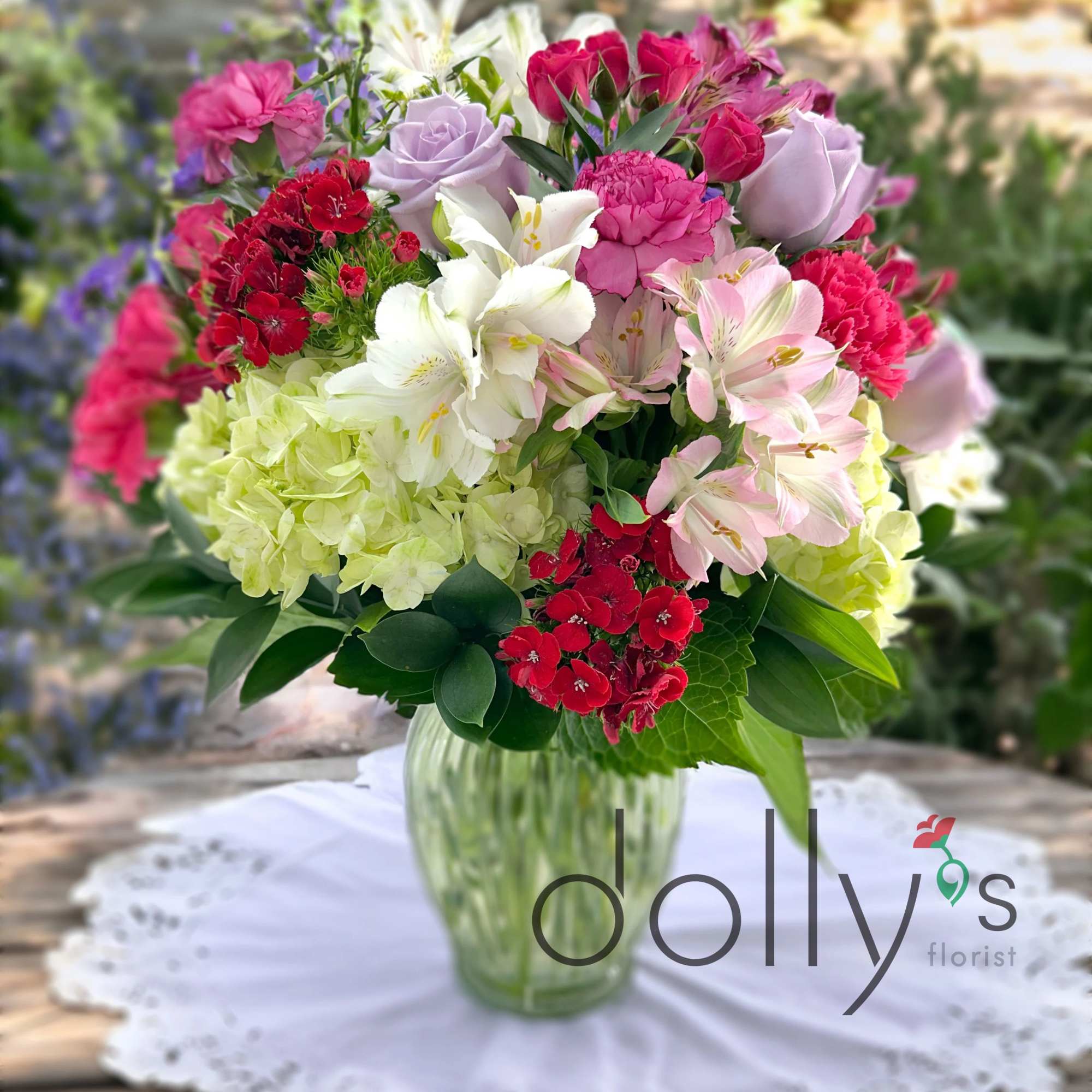 Mixed bouquet of pink, red, lavender, and white flowers in a green glass vase on a lace doily