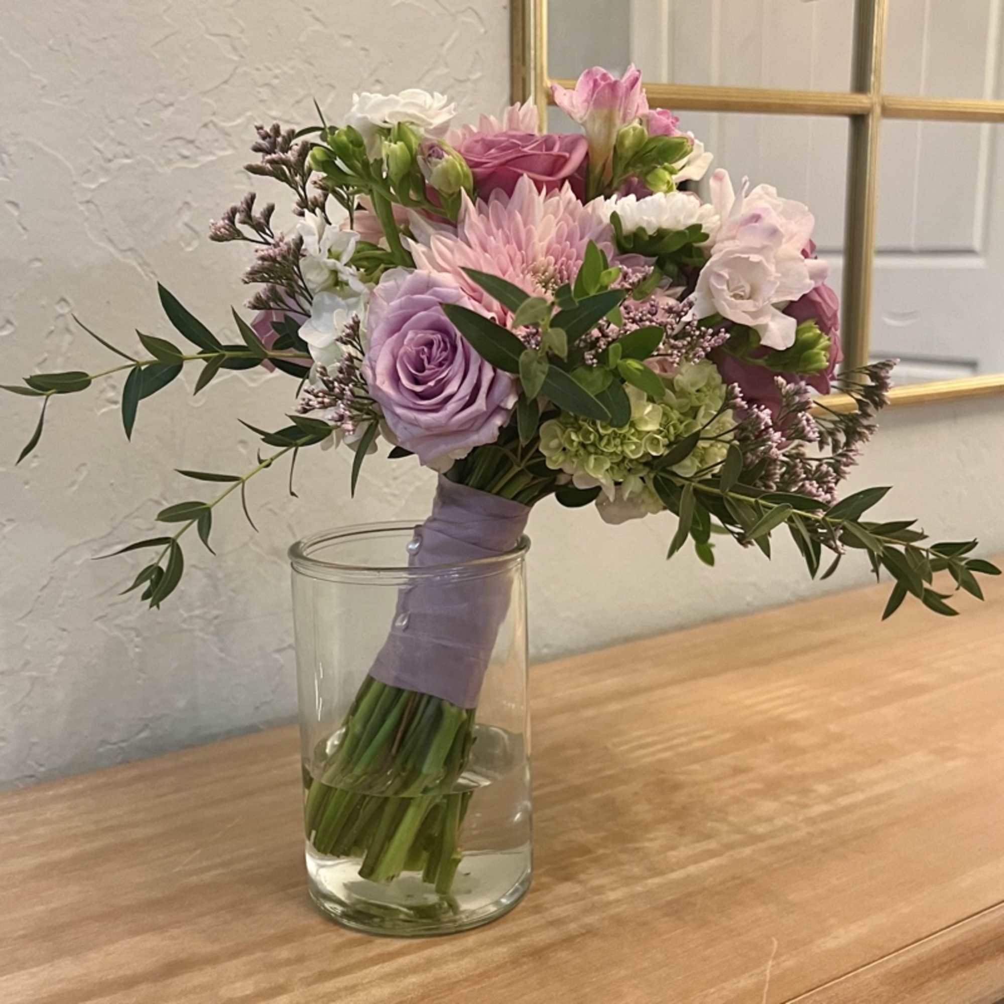 Hand-tied bouquet of lavender roses and pastel blooms in a clear glass vase on a wooden surface.