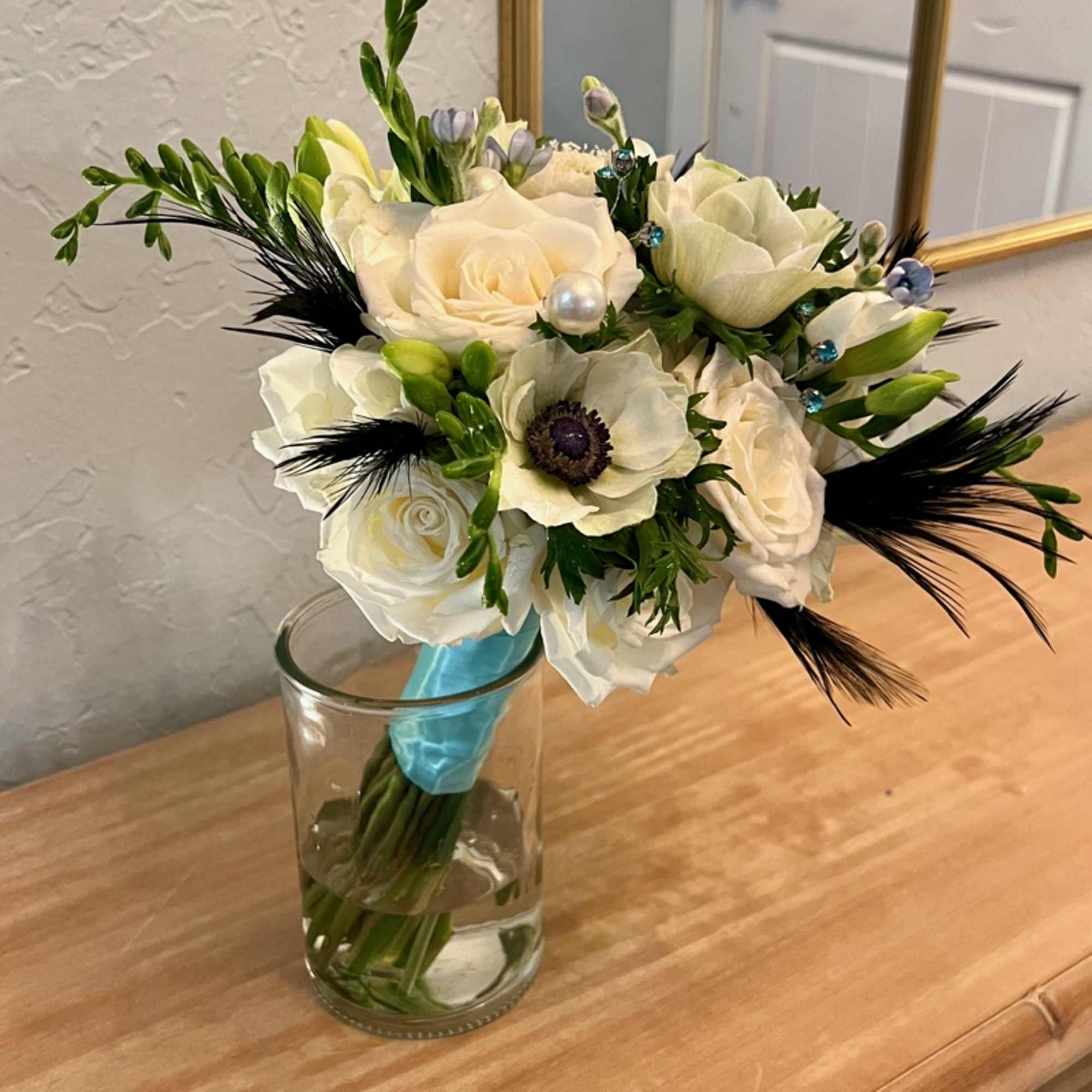 Hand-tied bouquet of white roses and anemones with black feathers in a clear glass vase.