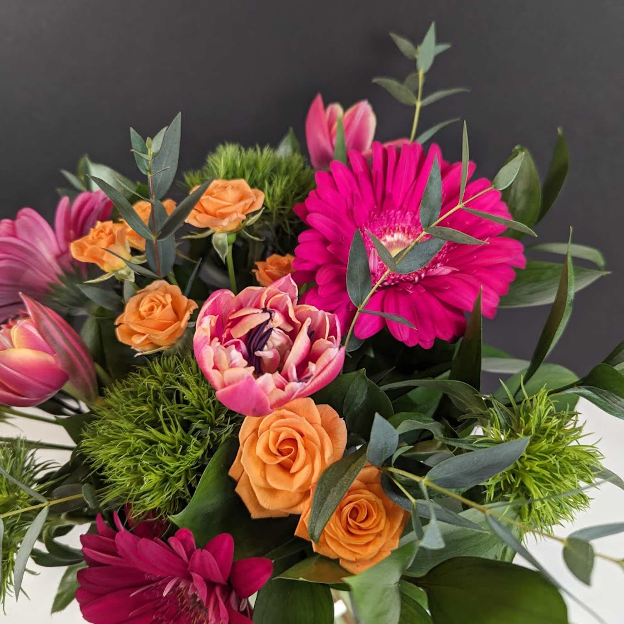 Bright bouquet of hot pink gerbera daisies, orange roses, and pink tulips in a clear glass vase.