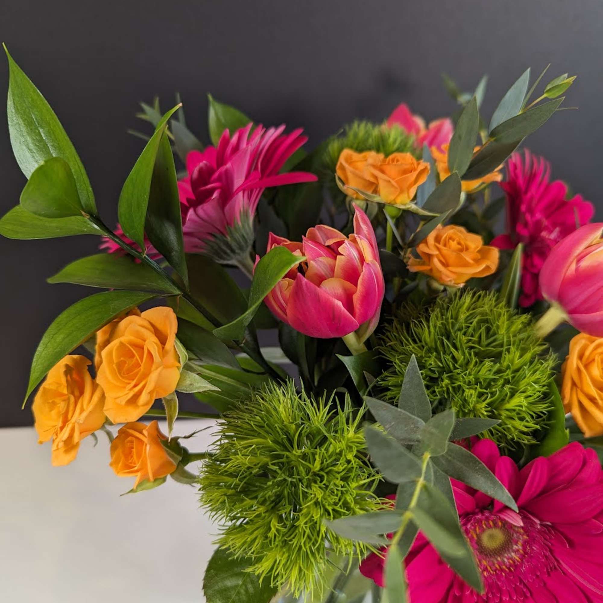Bright bouquet of hot pink gerbera daisies, orange spray roses, tulips and green dianthus in a clear glass vase