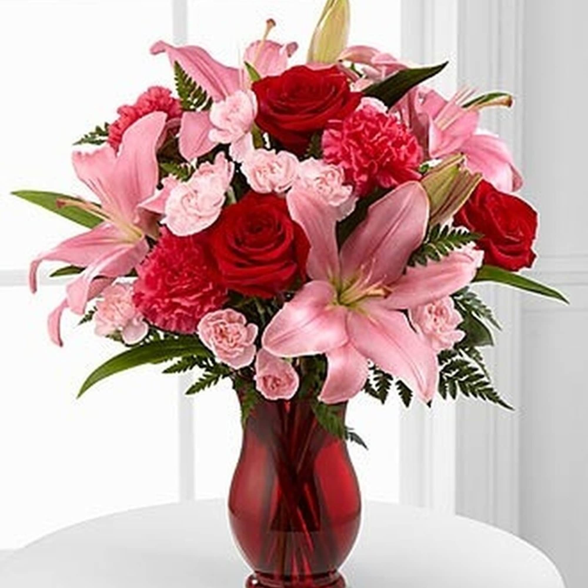 Arrangement of pink lilies, red roses, and pink carnations in a red glass vase