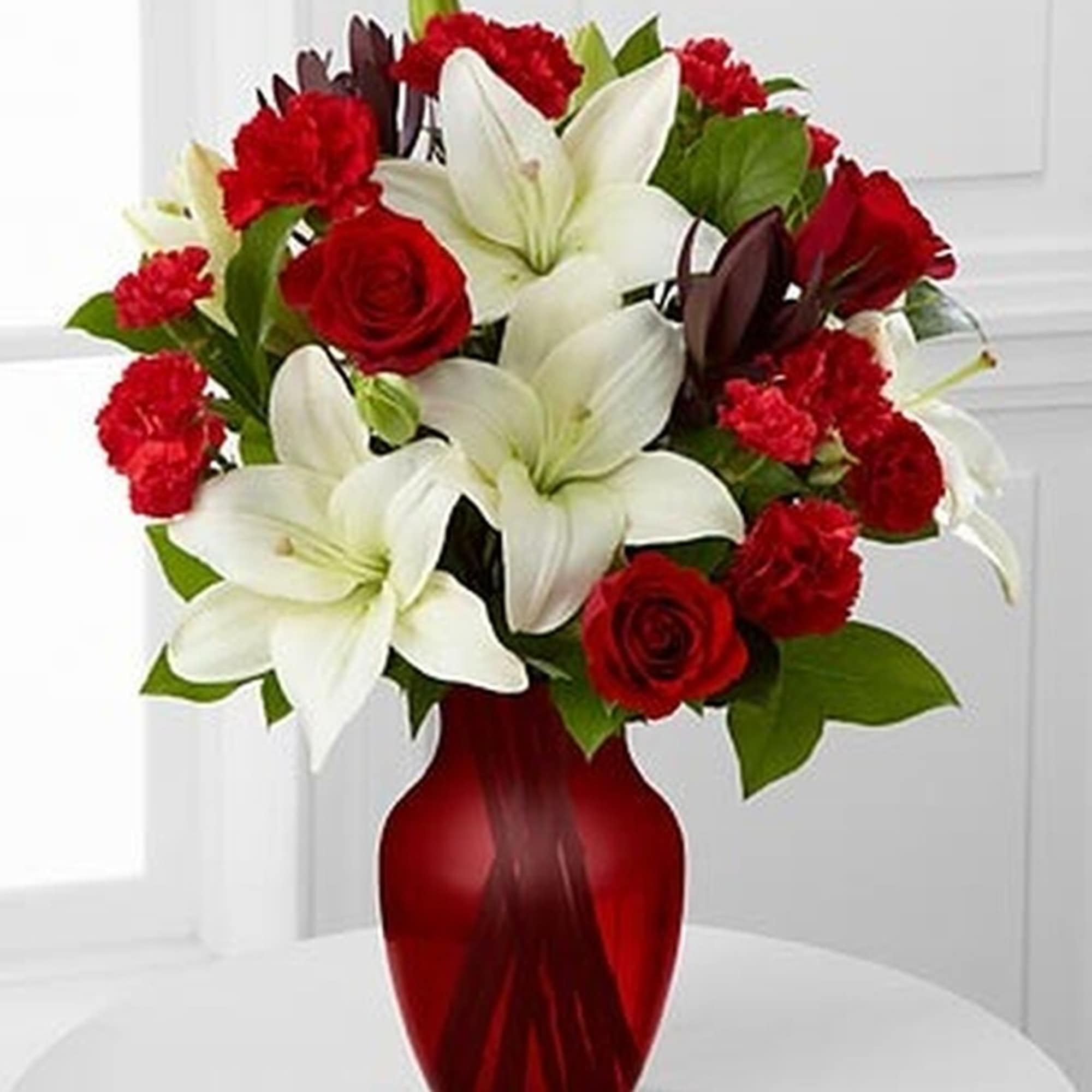 Arrangement of white lilies, red roses, and red carnations in a red glass vase.