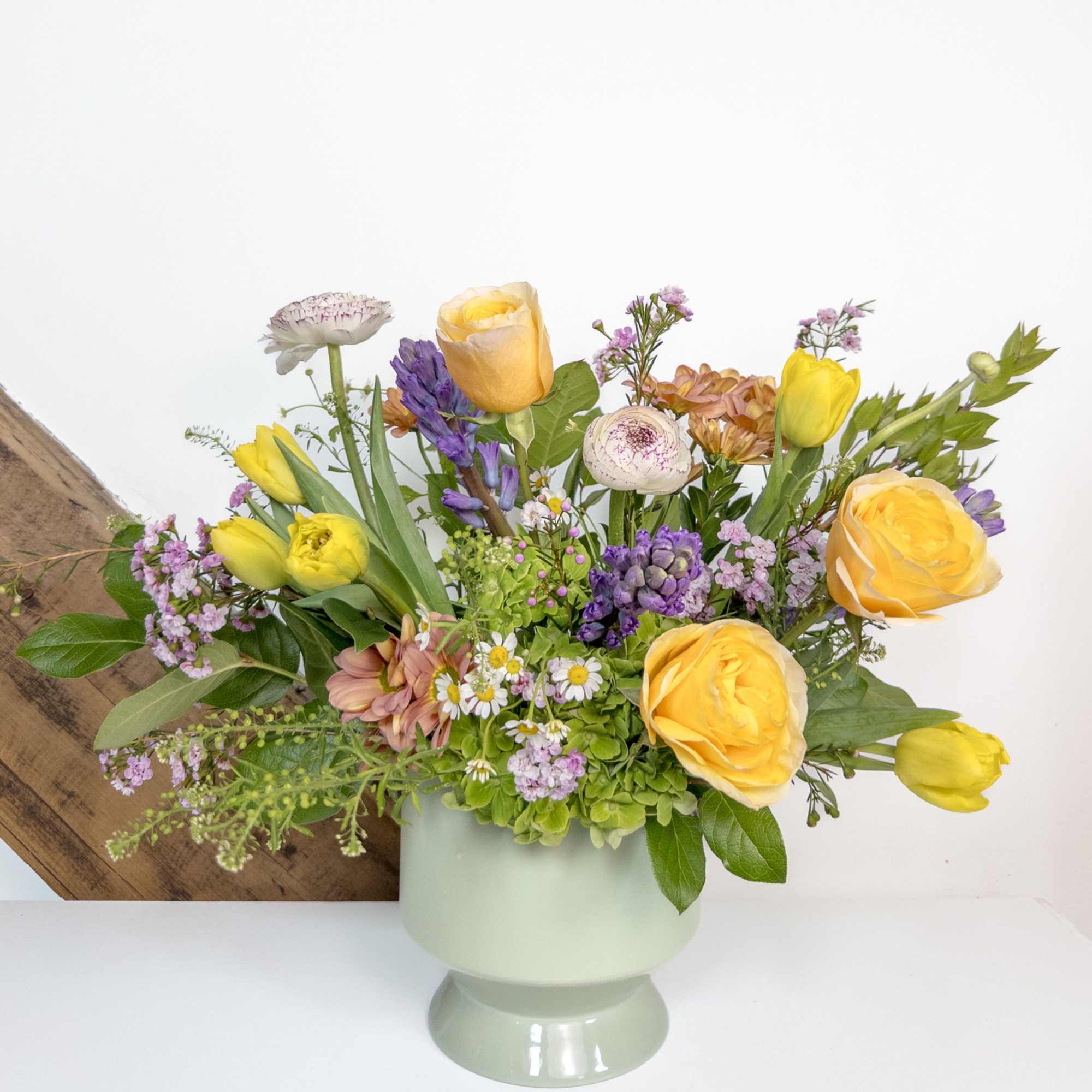 Low arrangement of yellow roses, tulips, and mixed pastel flowers in a pale green pedestal vase