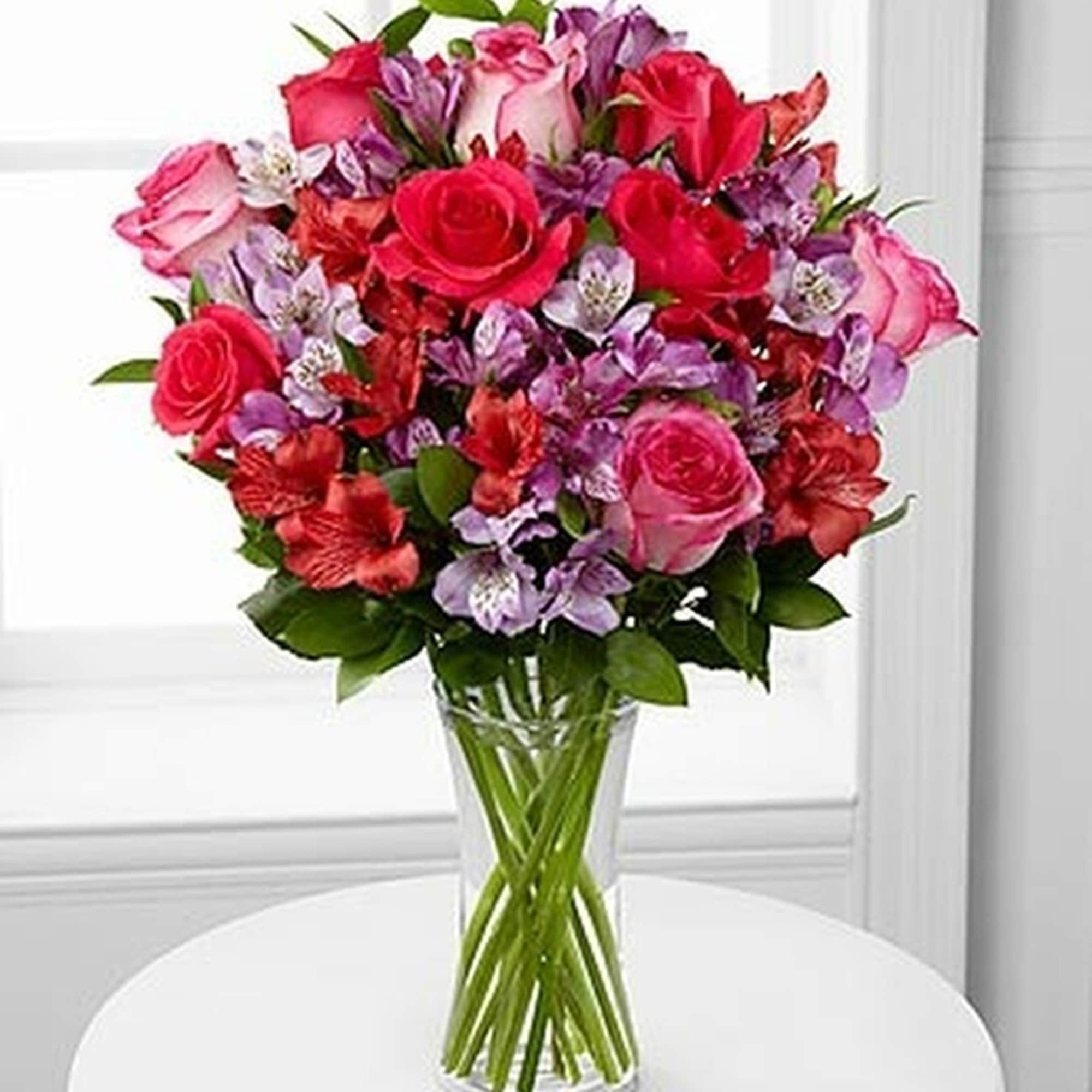 Tall glass vase of pink and red roses mixed with purple and white blooms