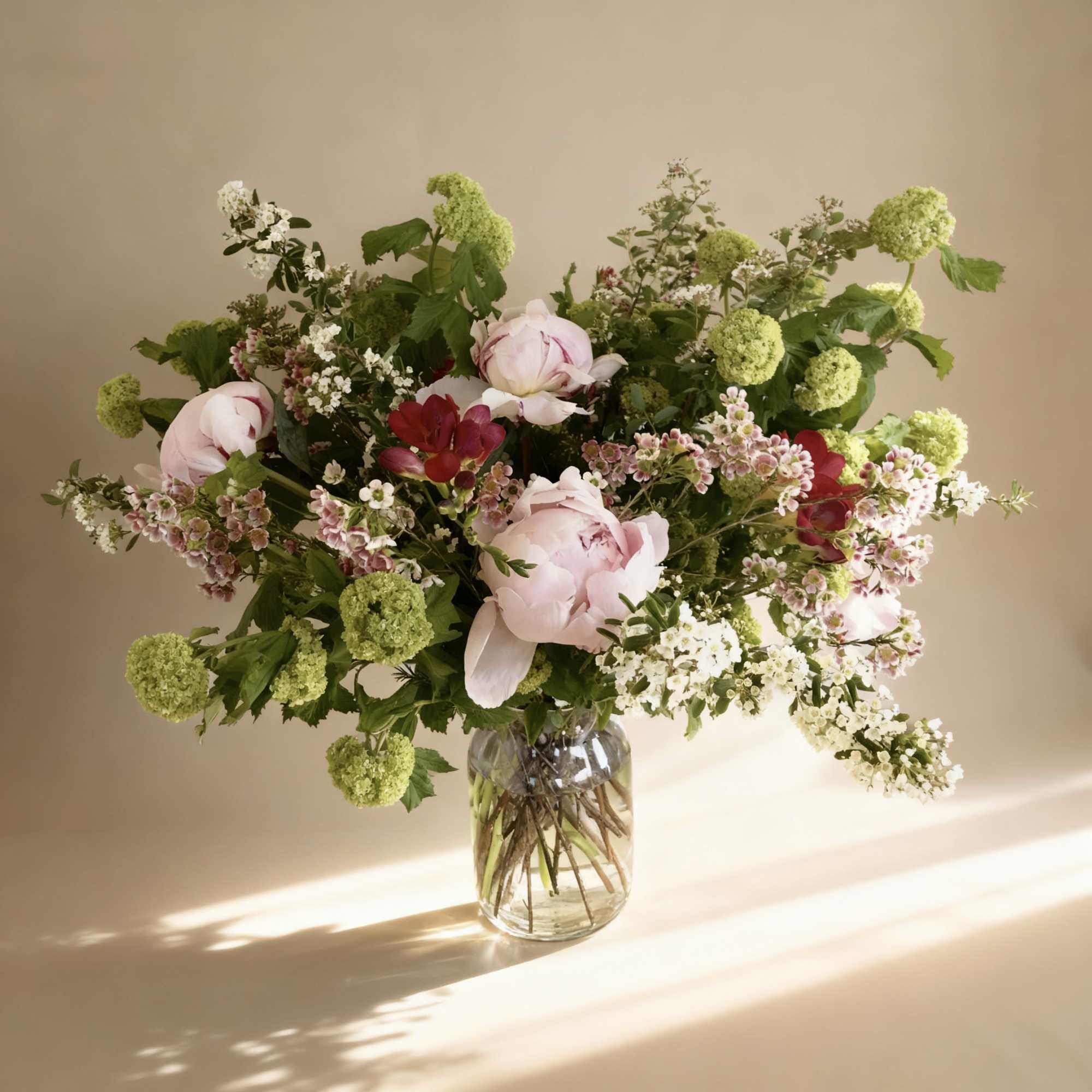 Loose arrangement of pale pink peonies, red blooms, and green flower clusters in a clear glass vase.