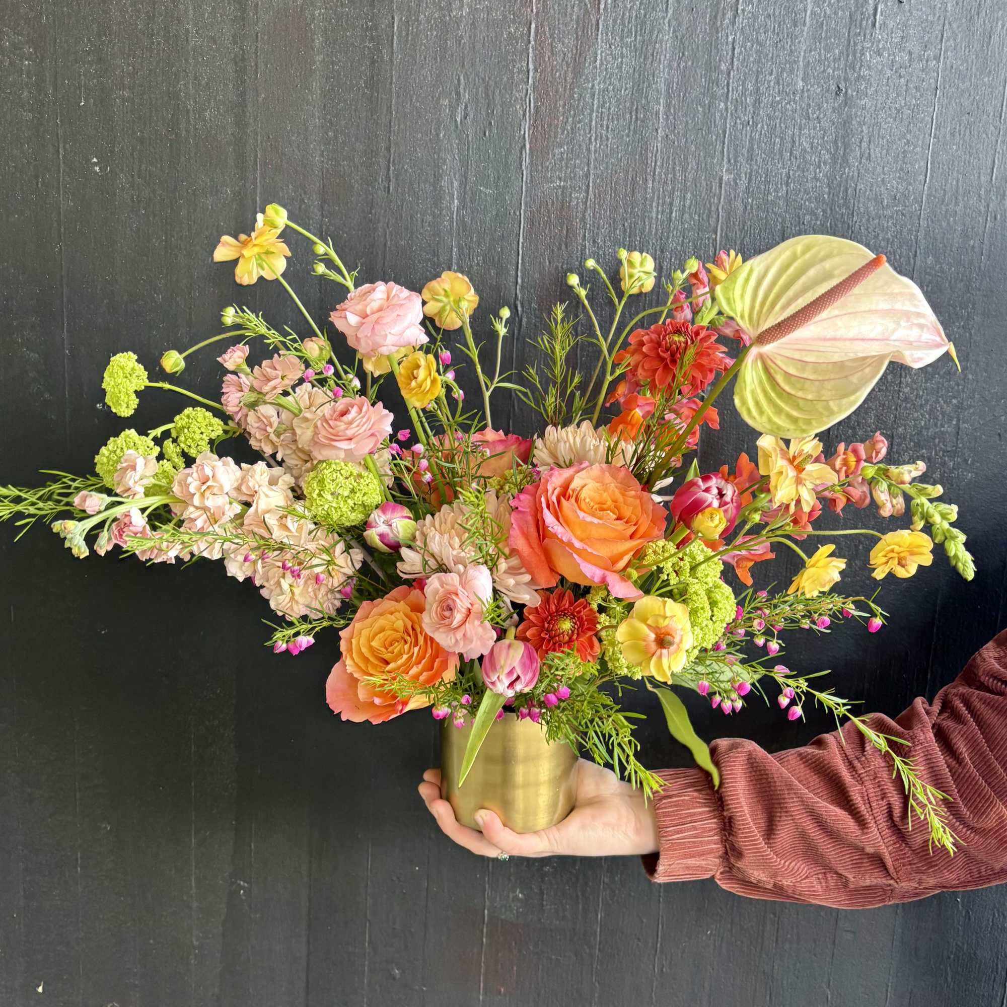 Colorful arrangement of pink, orange, and yellow flowers in a gold vase held against a dark background