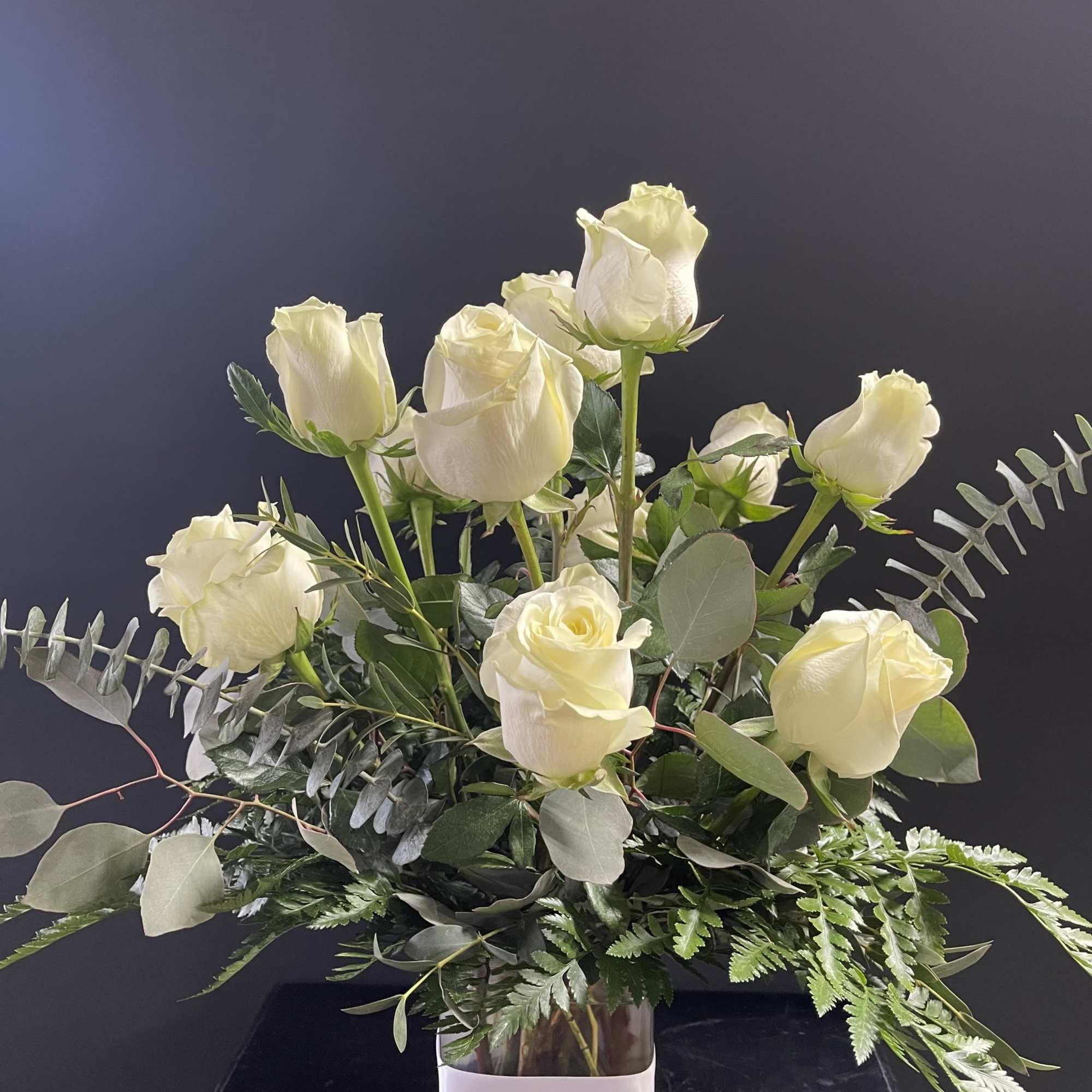 Arrangement of white roses in a clear square glass vase with foliage against a dark background